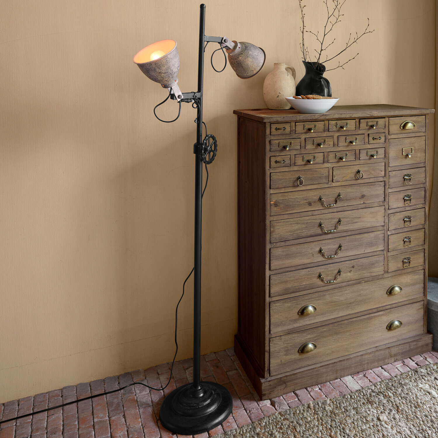 Lampada a stelo Fenric Lampada da terra con paralumi in metallo grigio e base nera accanto a una cassettiera in legno rustico