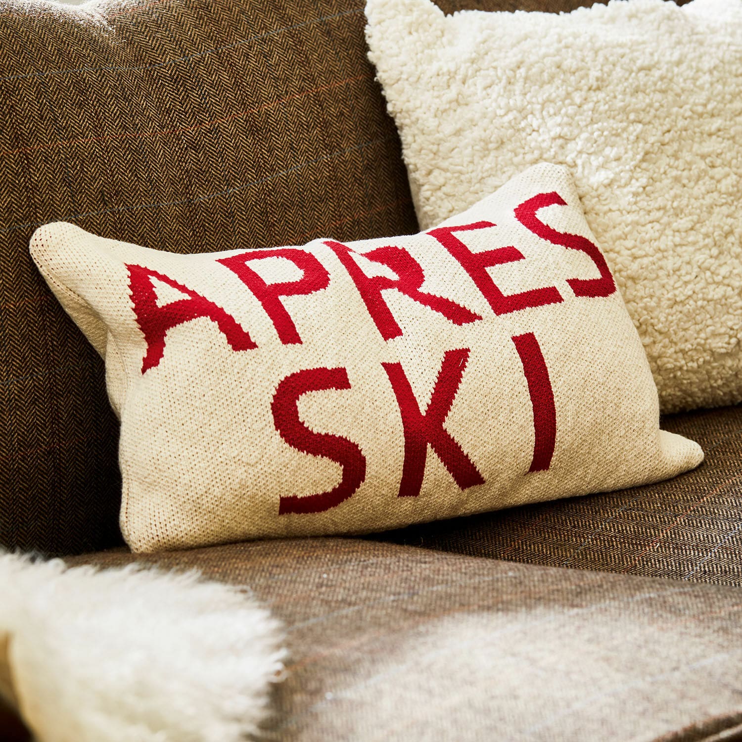 Copricuscino Apres Ski Cuscino beige con scritta rossa Apres Ski su divano marrone con coperta pelosa morbida