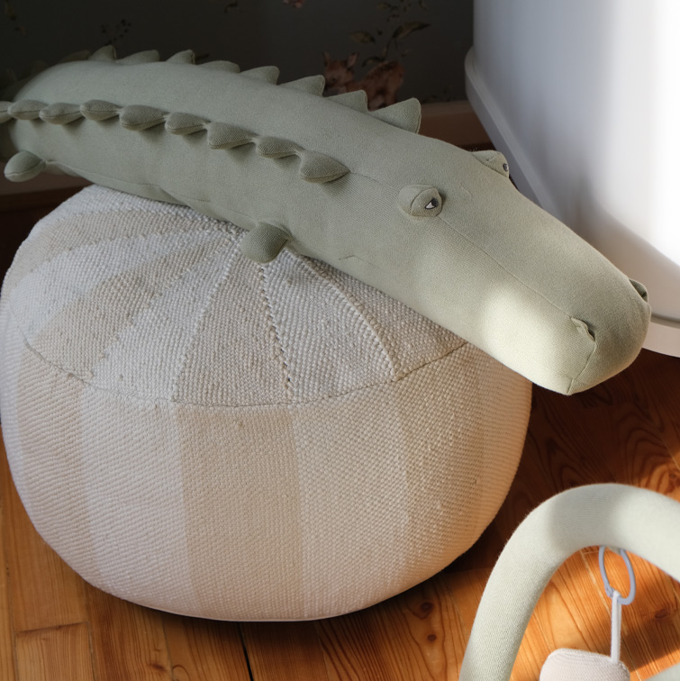 Coccodrillo di peluche verde chiaro su pouf beige intrecciato su pavimento in legno, decorazione per bambini