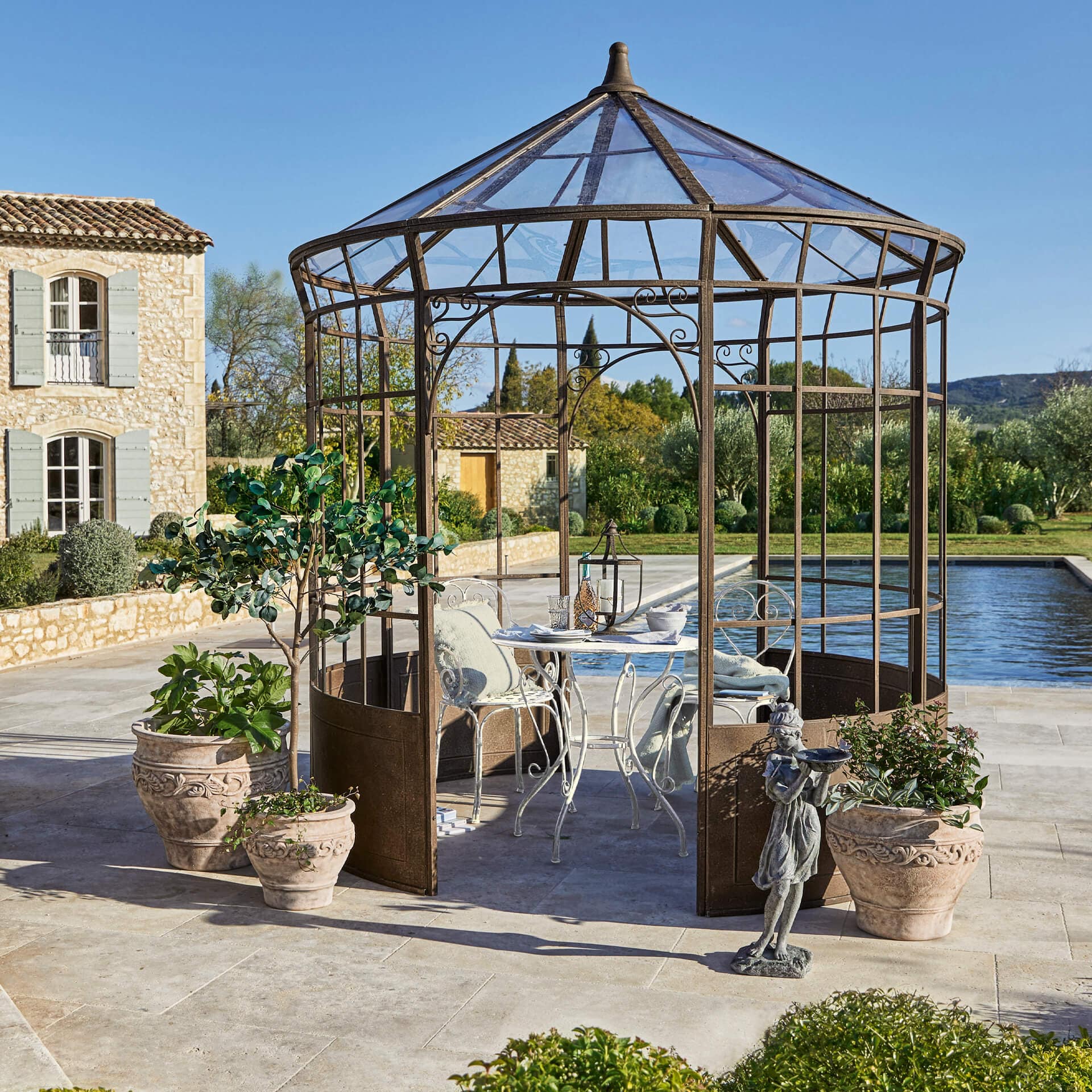 Giardino con gazebo in metallo ruggine, tavolo e sedie bianche, circondato da vasi con piante