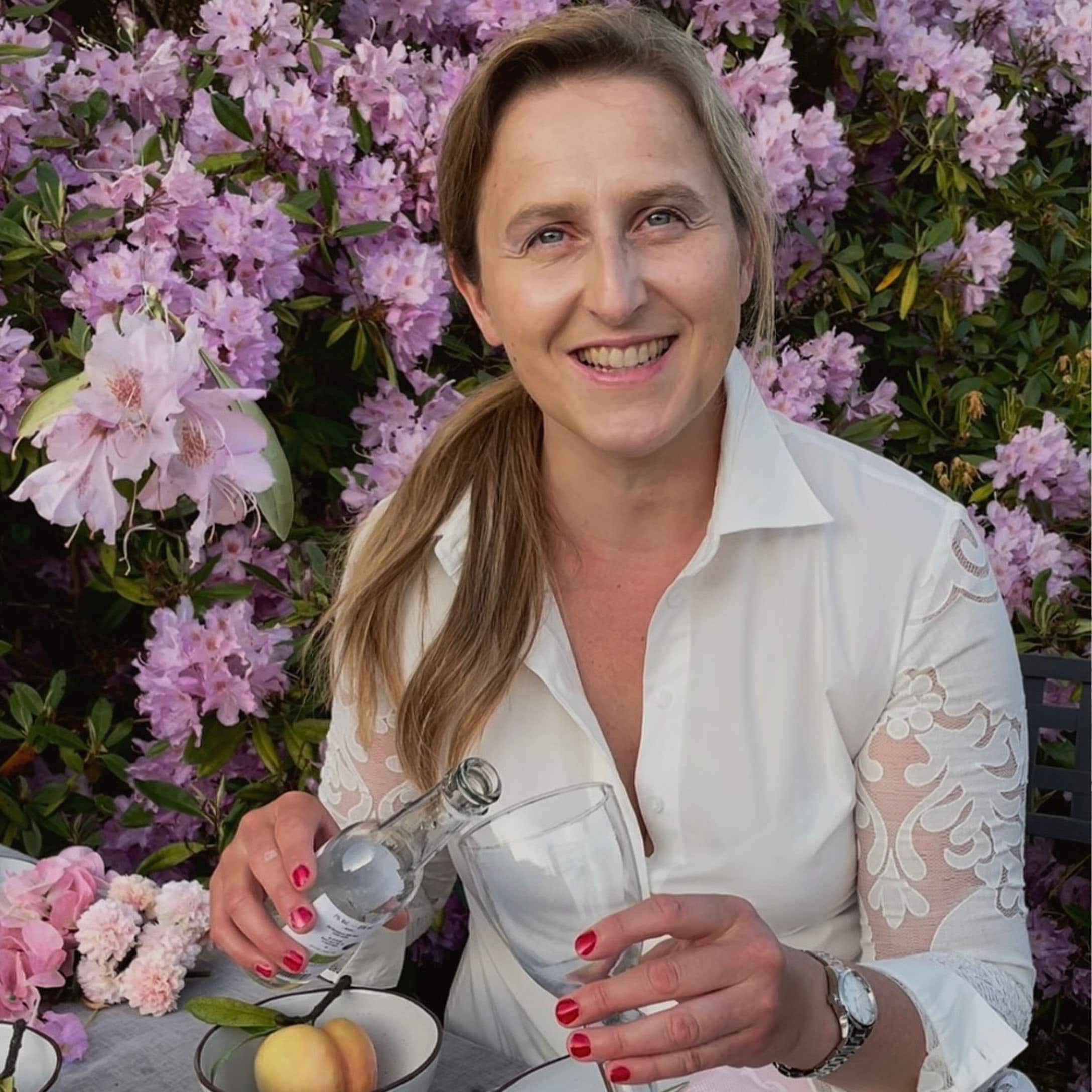 Donna con camicia bianca e maniche di pizzo versa bevanda in bicchiere davanti a fiori rosa