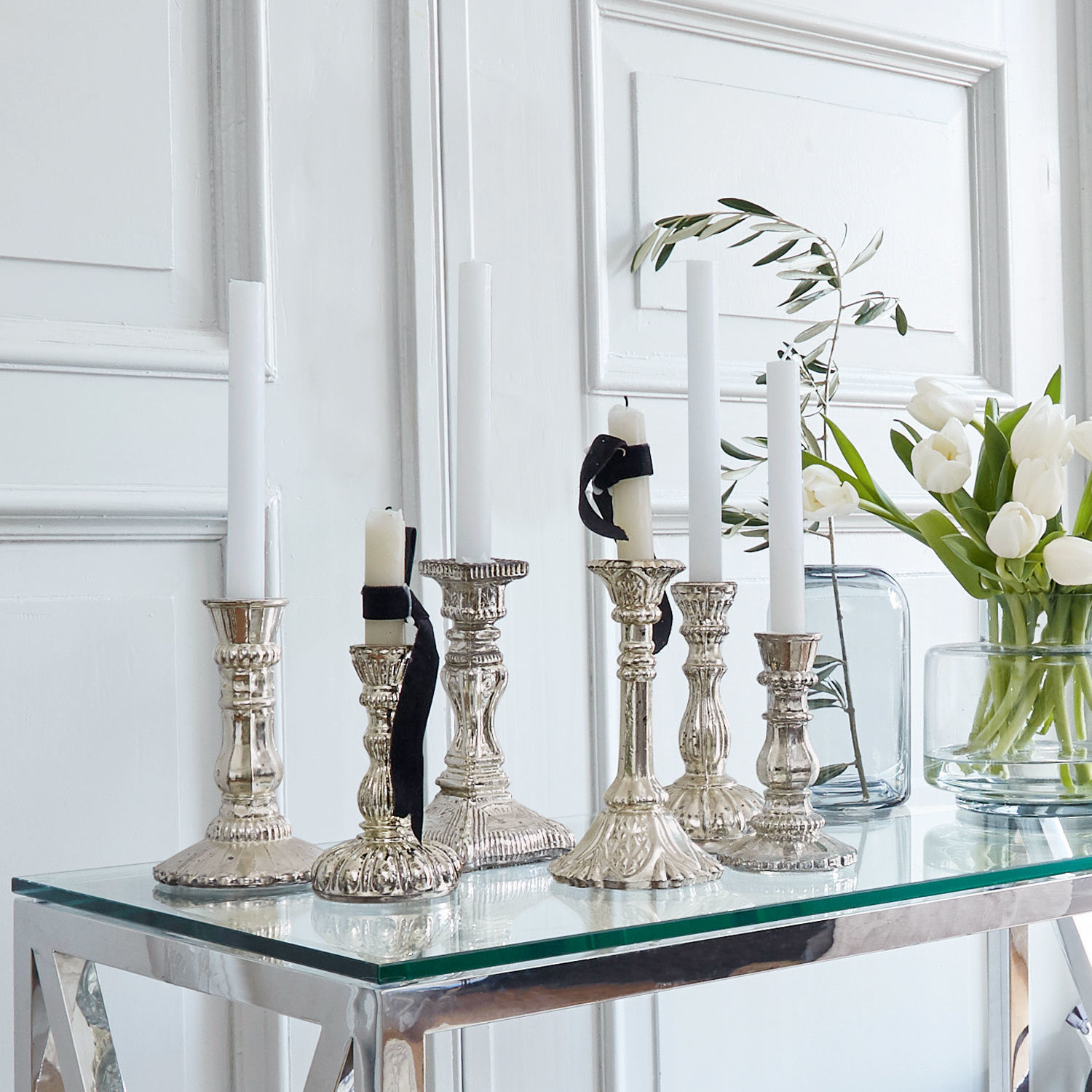Set di 6 portacandele Adolpha Set di candelabri in metallo argentato con candele bianche su tavolo in vetro in interno luminoso