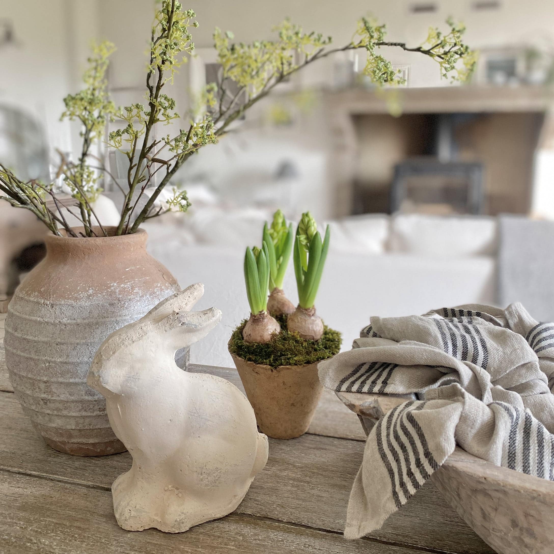 Coniglio decorativo bianco in ceramica accanto a vaso con germogli verdi e tessuto di lino su tavolo in legno