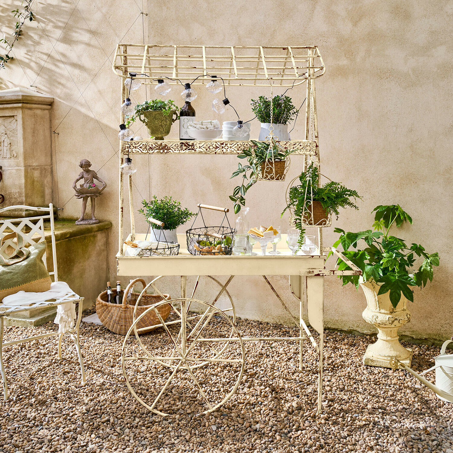 Carrello bar Gavrielle Carrello da giardino in metallo crema con piante e decorazioni su patio di ghiaia