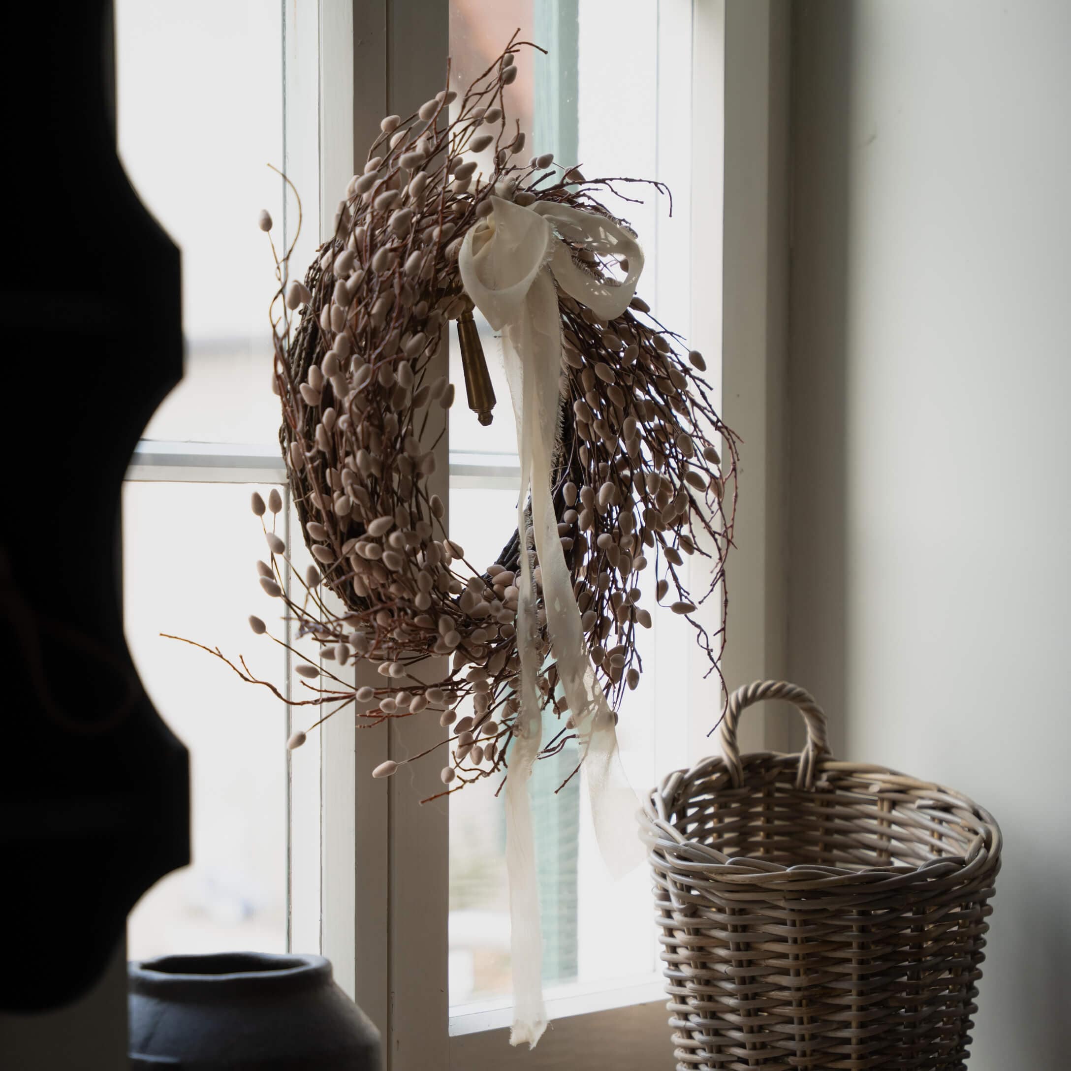 Corona decorativa beige di rami e boccioli con nastro bianco appesa alla finestra, cesto di vimini accanto