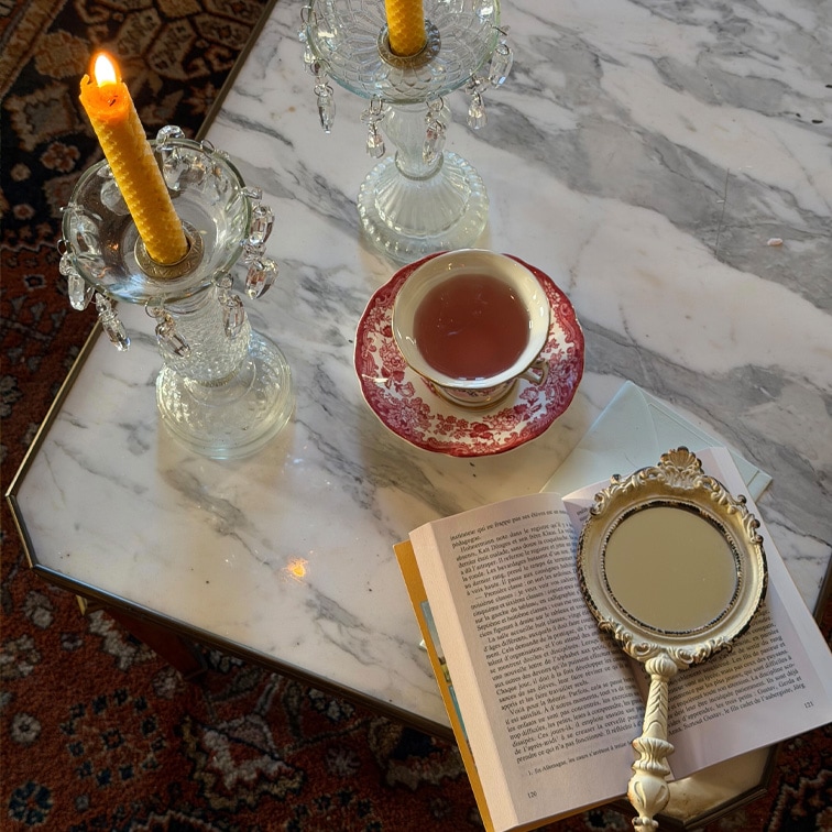 Candelabri in vetro con candele gialle e tazza con motivo rosso su tavolo in marmo con libro e specchio a mano