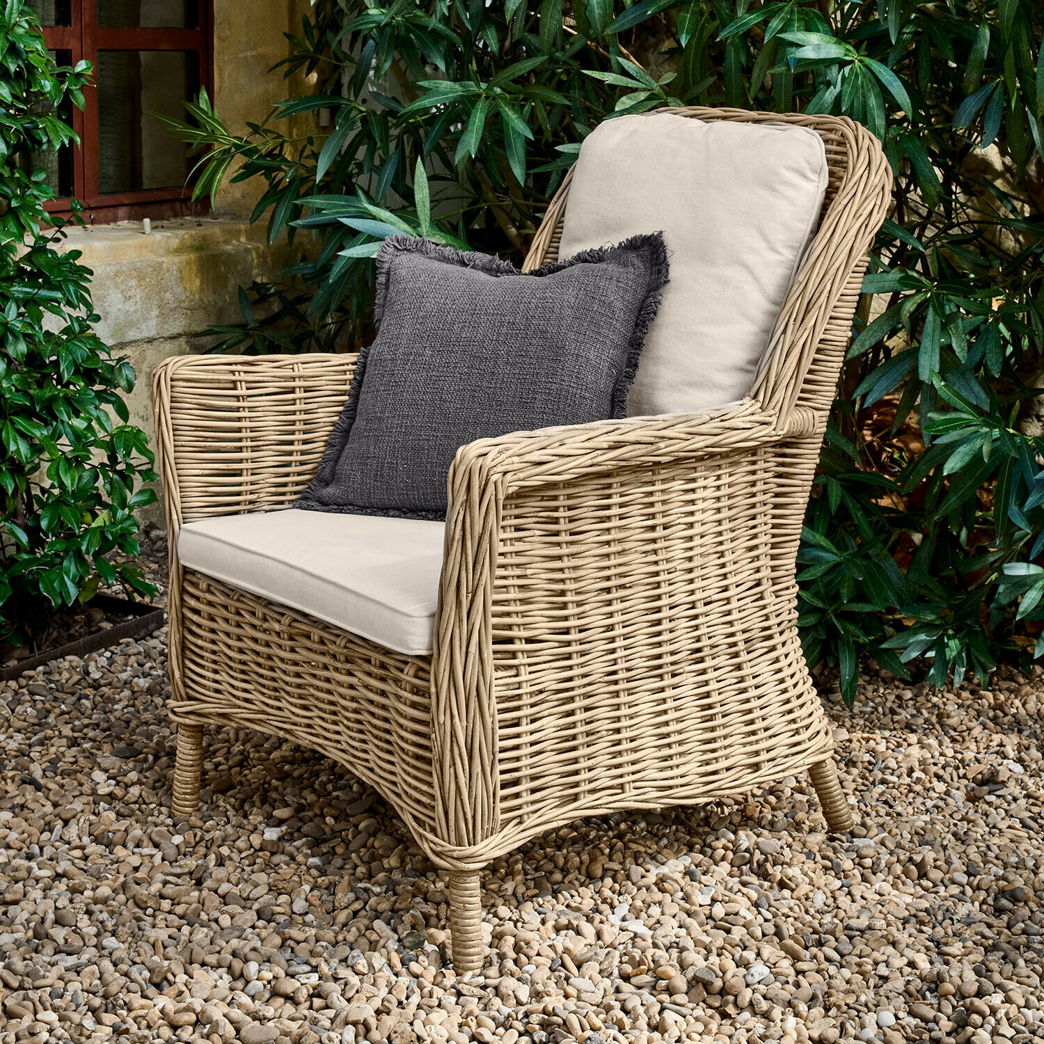 Poltrona Travenne Poltrona in rattan con cuscini crema e cuscino decorativo grigio scuro su patio di ghiaia