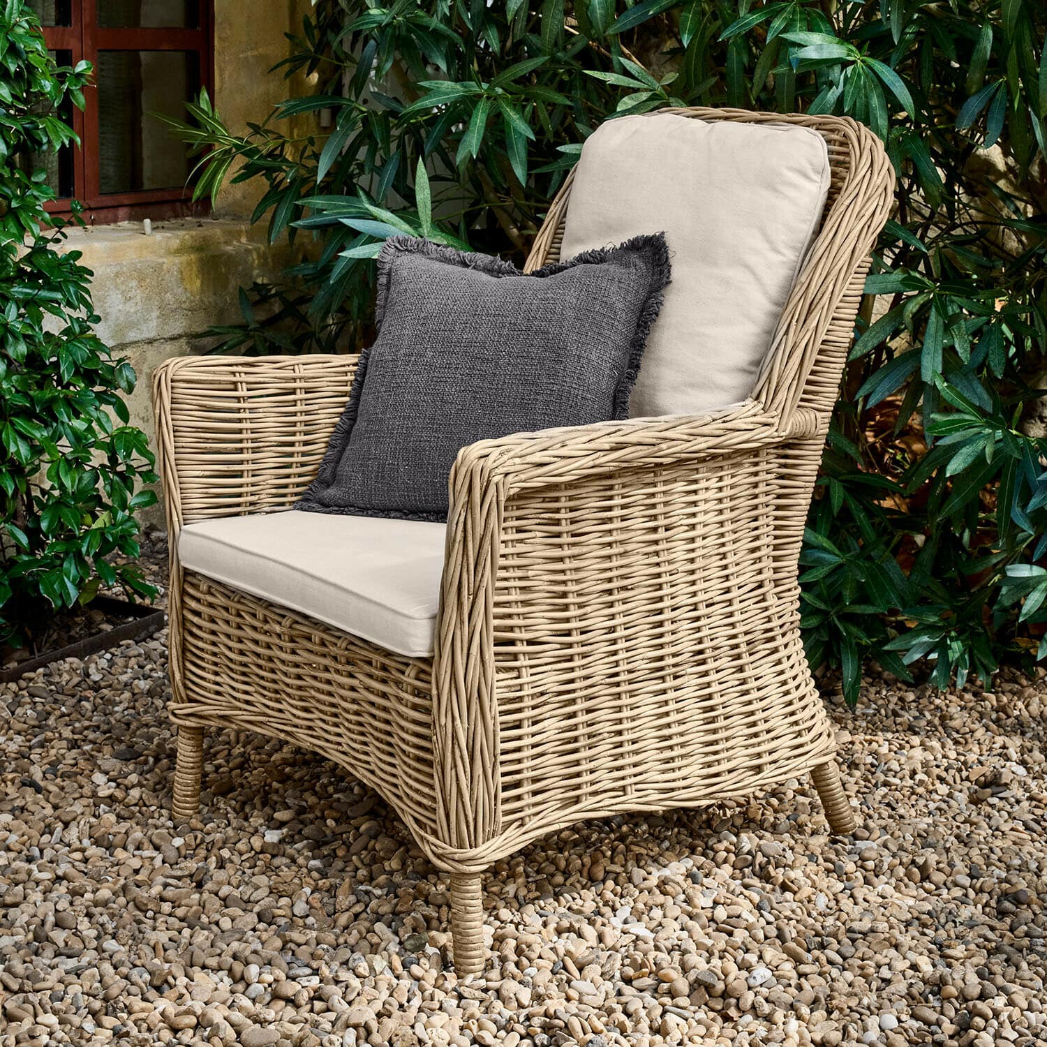 Poltrona Travenne Poltrona in rattan con cuscini crema e cuscino decorativo grigio scuro su patio di ghiaia