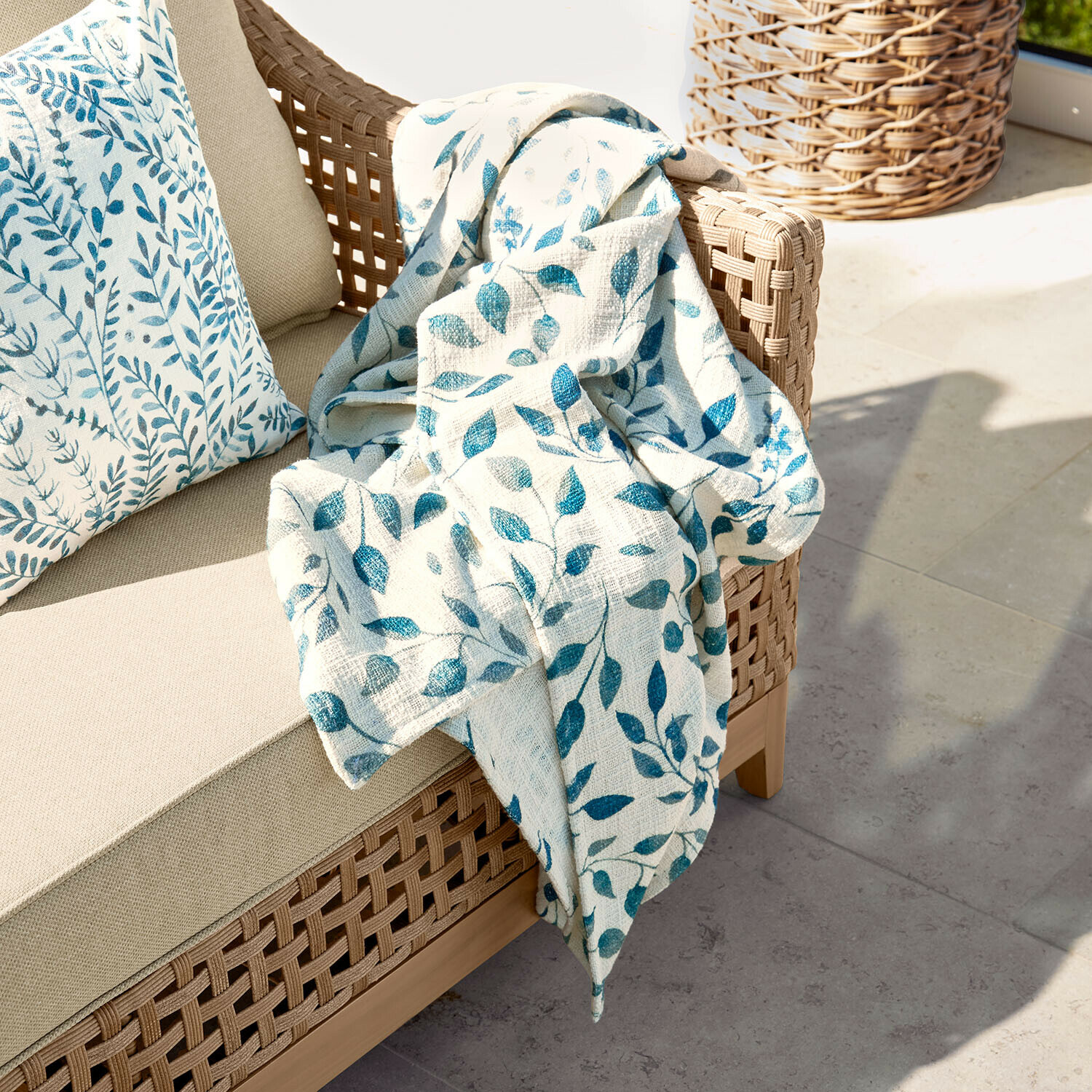 Coperta Ocevara Divano beige con schienale intrecciato e coperta crema con motivo foglie blu su terrazza