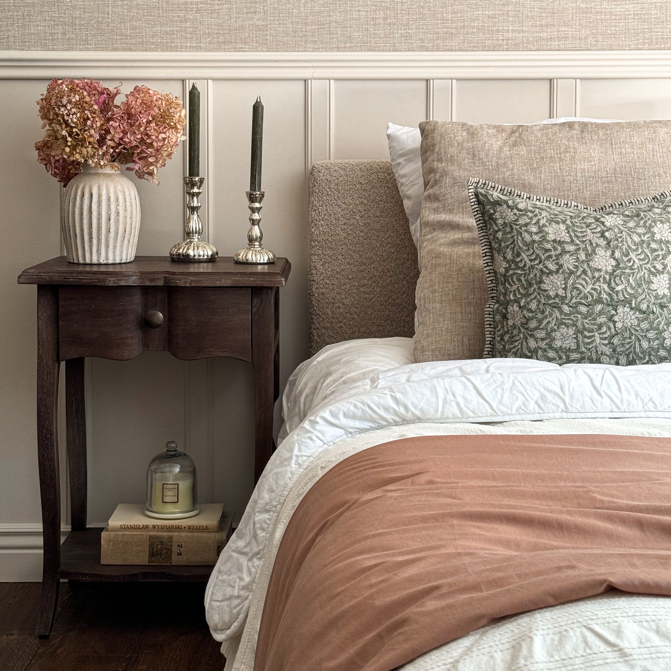Camera da letto accogliente con biancheria beige, cuscino verde e comodino in legno con decorazioni