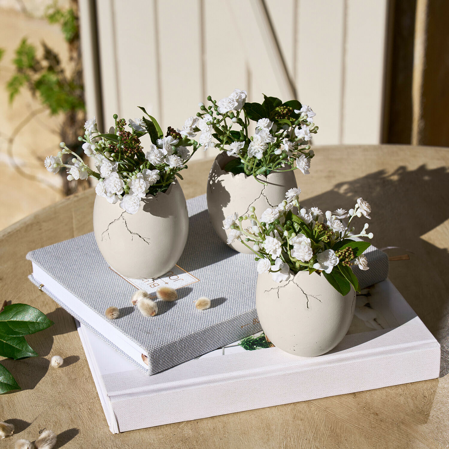 Set di 3 uova decorative Waverlye Tre vasi in ceramica bianca a forma di uovo con fiori bianchi su tavolo di legno con libri