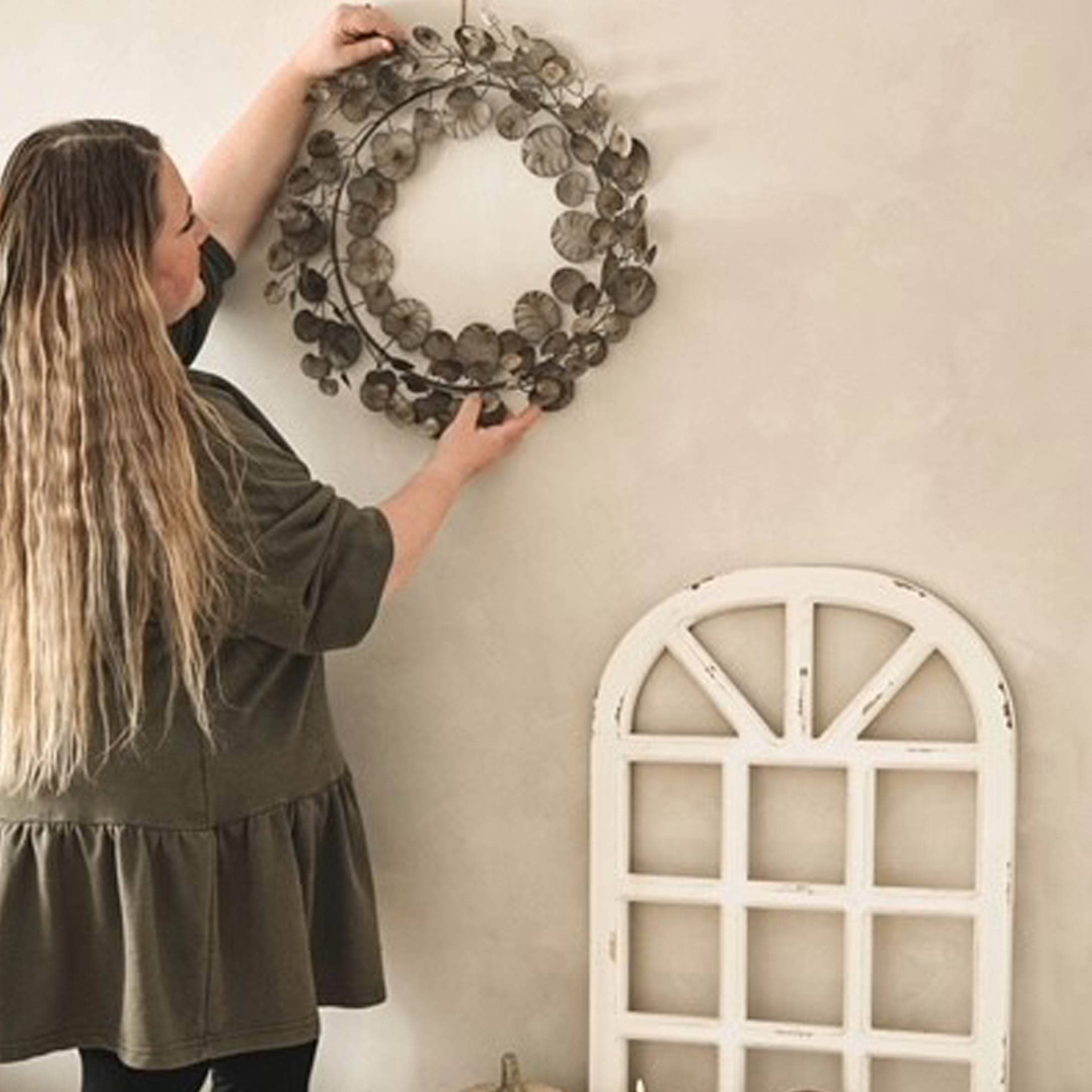 Donna che appende corona decorativa in metallo marrone su parete chiara in stile rustico