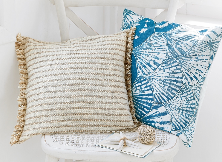 Due cuscini decorativi su sedia bianca, uno beige a righe con frange, l'altro blu con motivo conchiglia