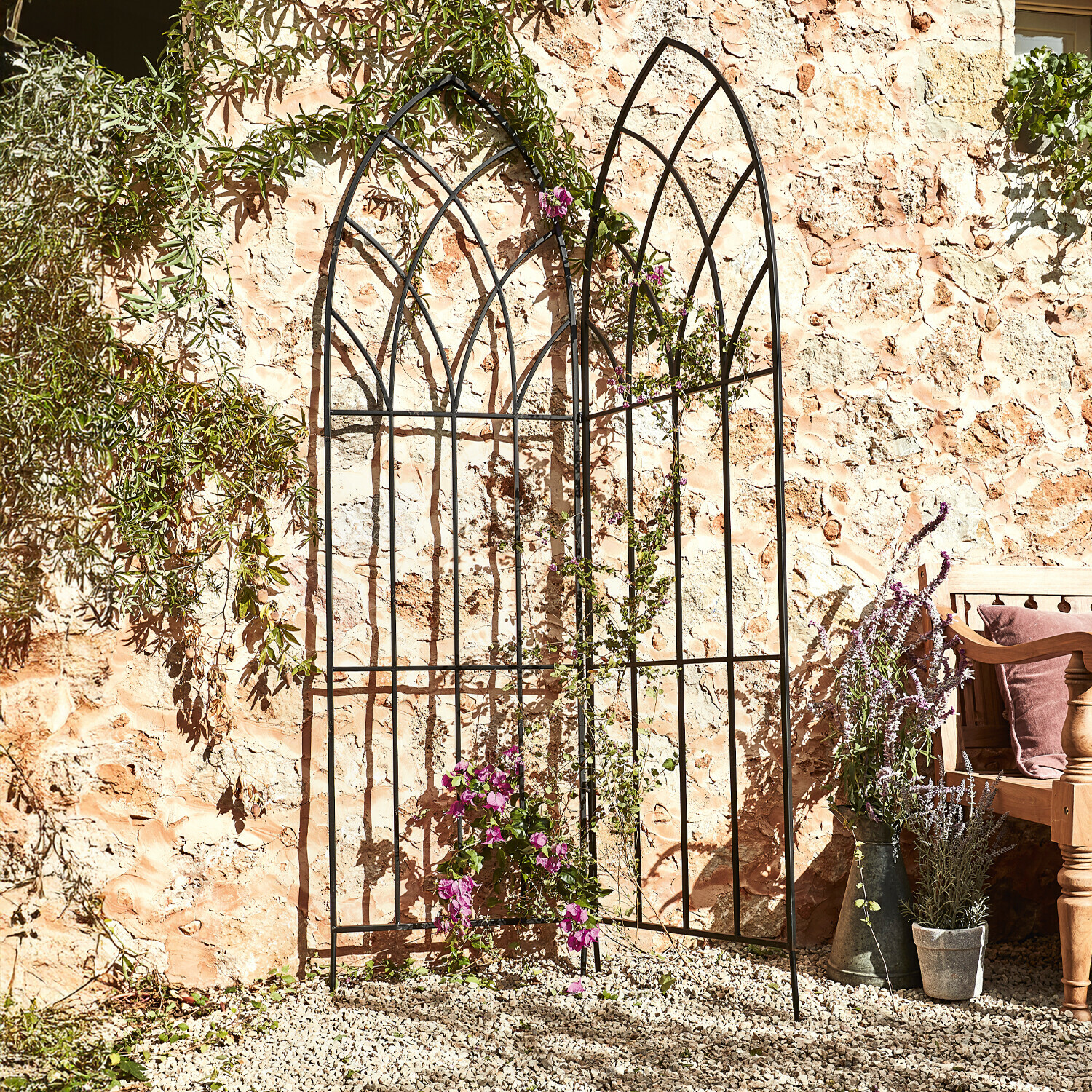 Traliccio per rampicanti Ophélie Grigliato da giardino in metallo nero con motivo ad arco davanti a muro in pietra con piante rampicanti