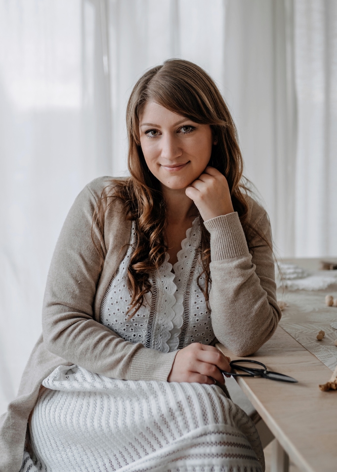 Donna con cardigan beige e vestito bianco in pizzo seduta a un tavolo di legno in ambiente luminoso