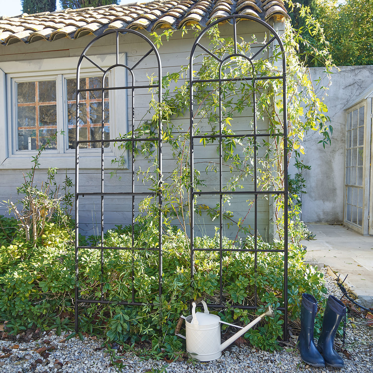 Coppia di tralicci per rampicanti Ealdred Griglia da giardino in metallo nero con motivo ad arco davanti a piante e casa, annaffiatoio e stivali blu