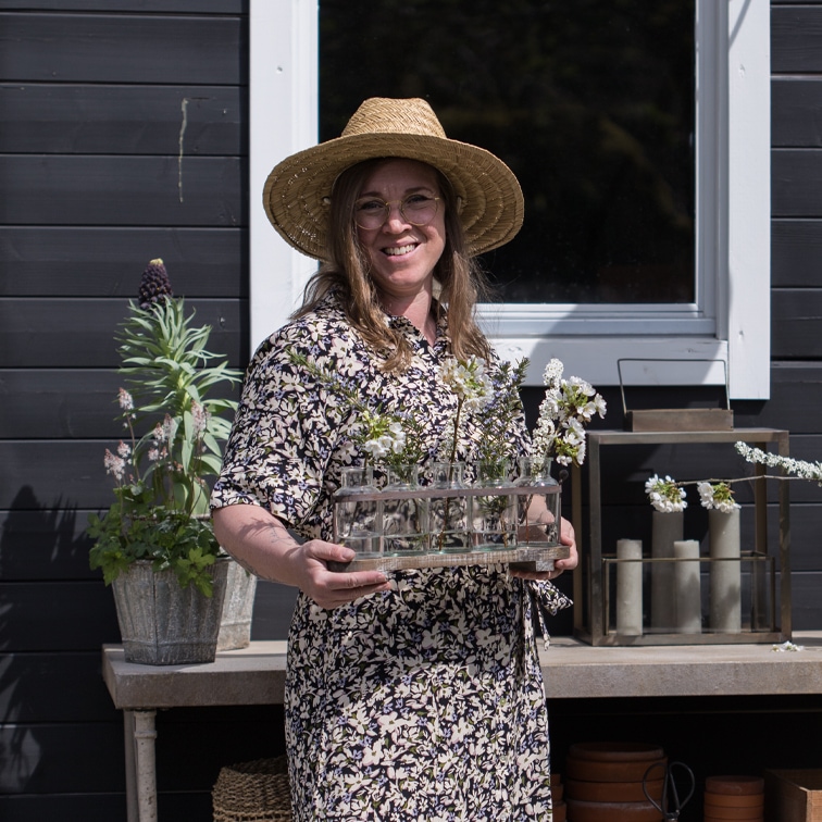 Donna con cappello che tiene decorazione con provette di vetro e fiori davanti a muro nero