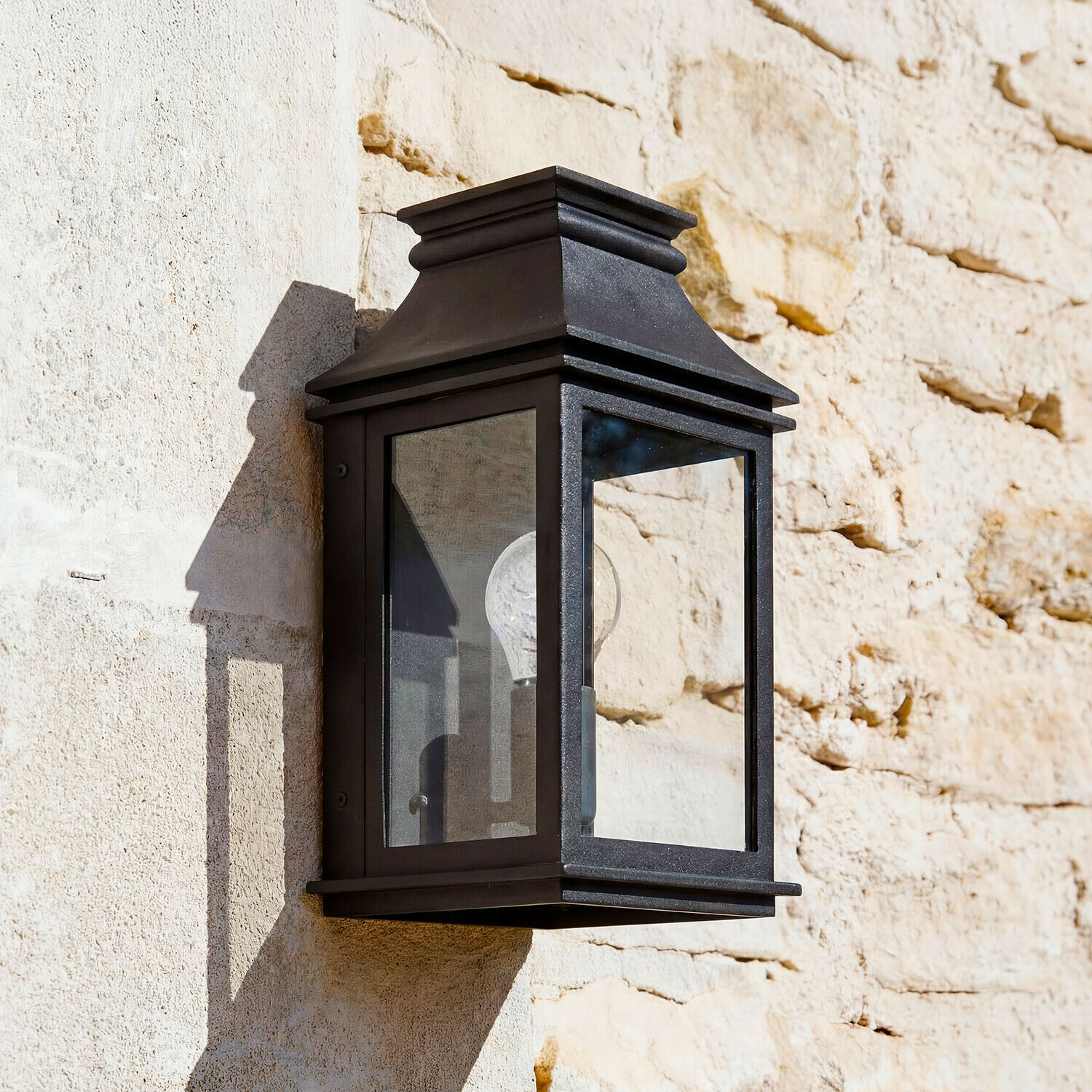 Lampada da parete per esterni Zélivarne Applique da parete nera in metallo con vetro trasparente su muro di pietra chiara, illuminazione esterna