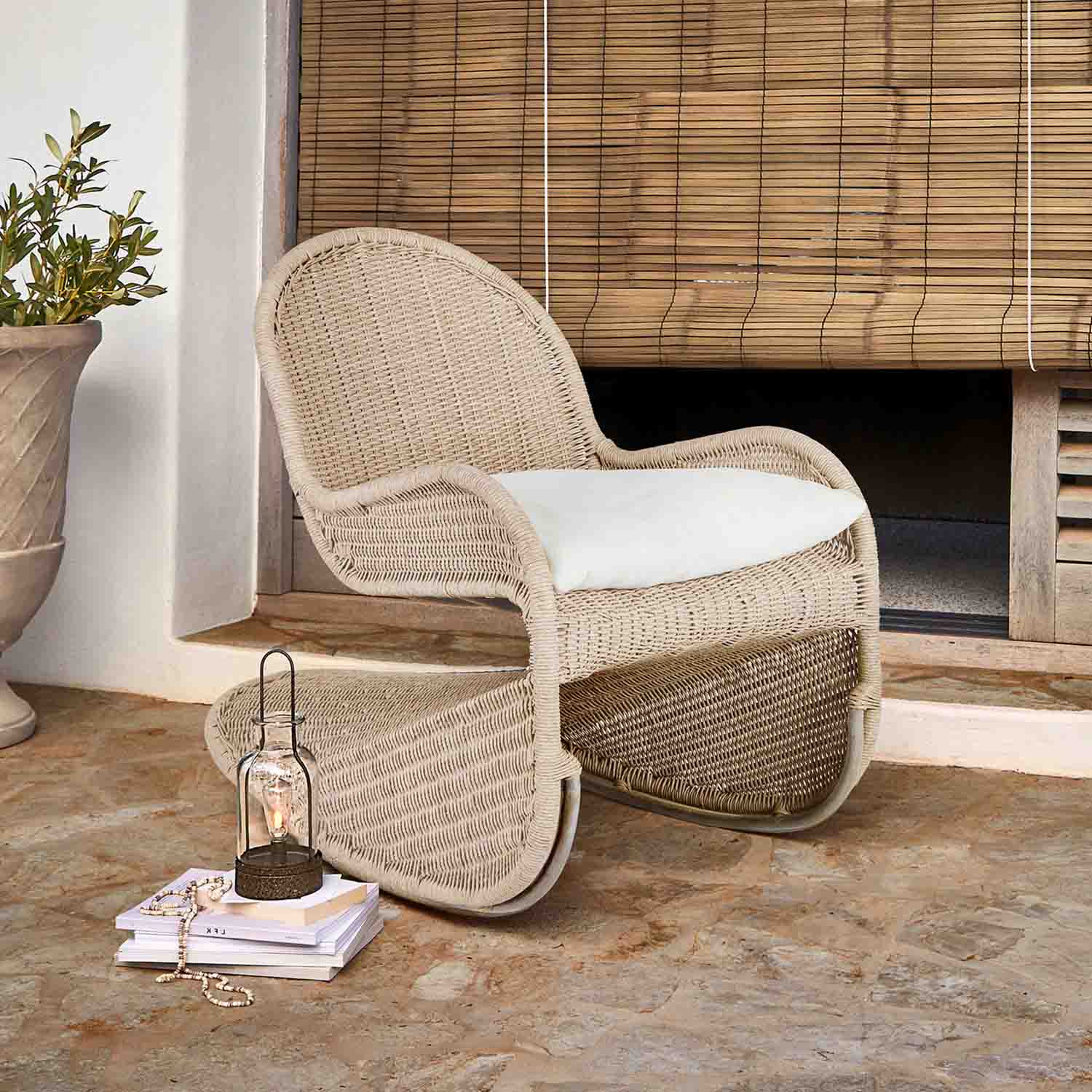 Poltrona in vimini beige con cuscino bianco su terrazza, accanto lanterna e libri su pavimento in pietra