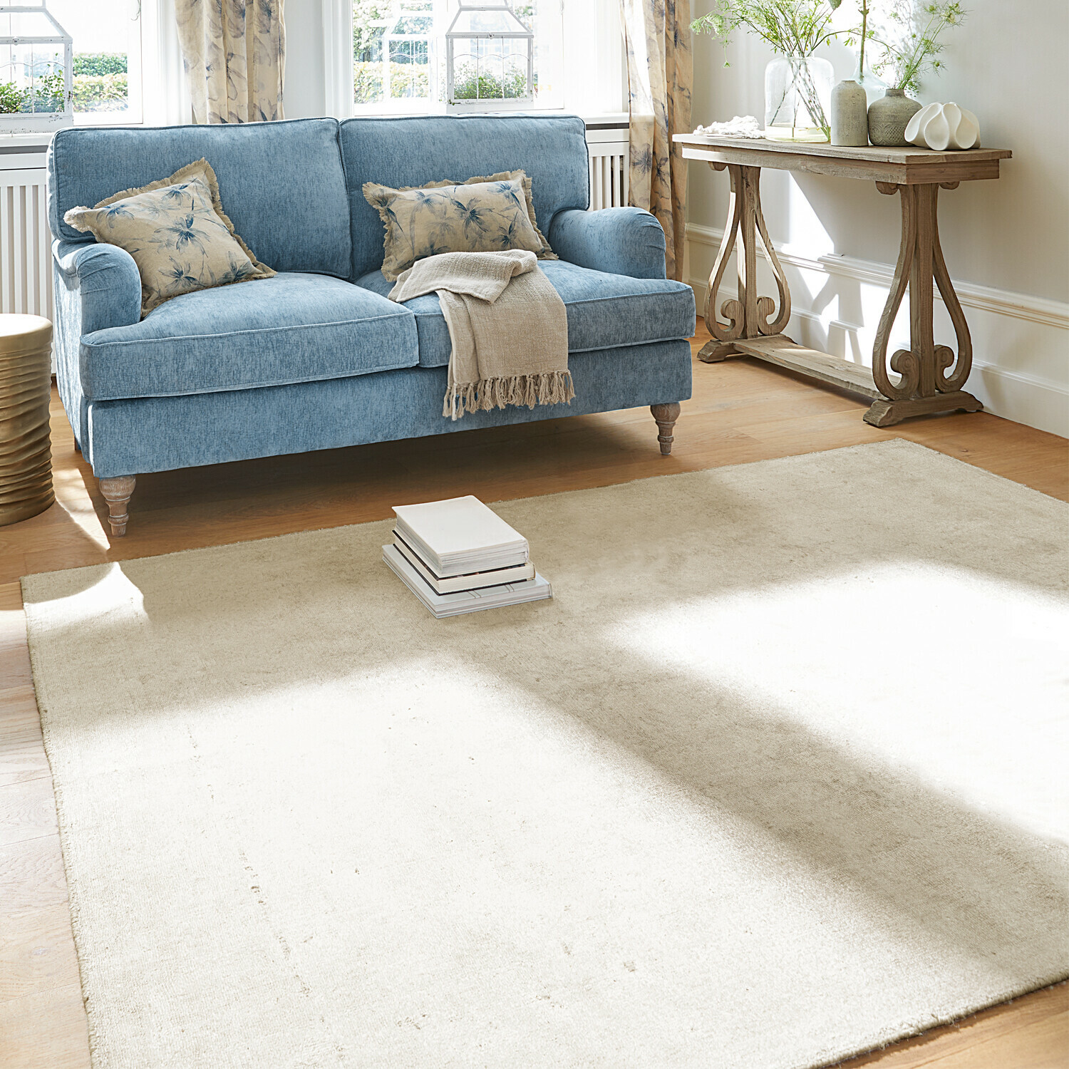 Tappeto Lorémont Divano blu in tessuto con coperta beige e cuscini, tappeto chiaro su pavimento in legno in soggiorno luminoso