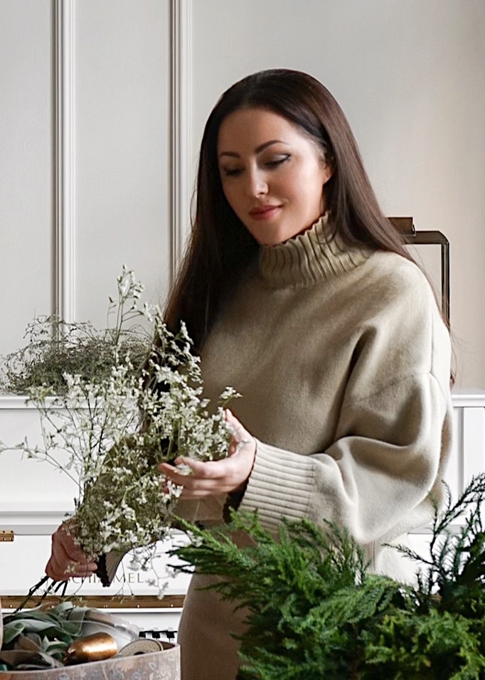 Donna con maglione beige a collo alto che tiene fiori bianchi delicati in interno luminoso