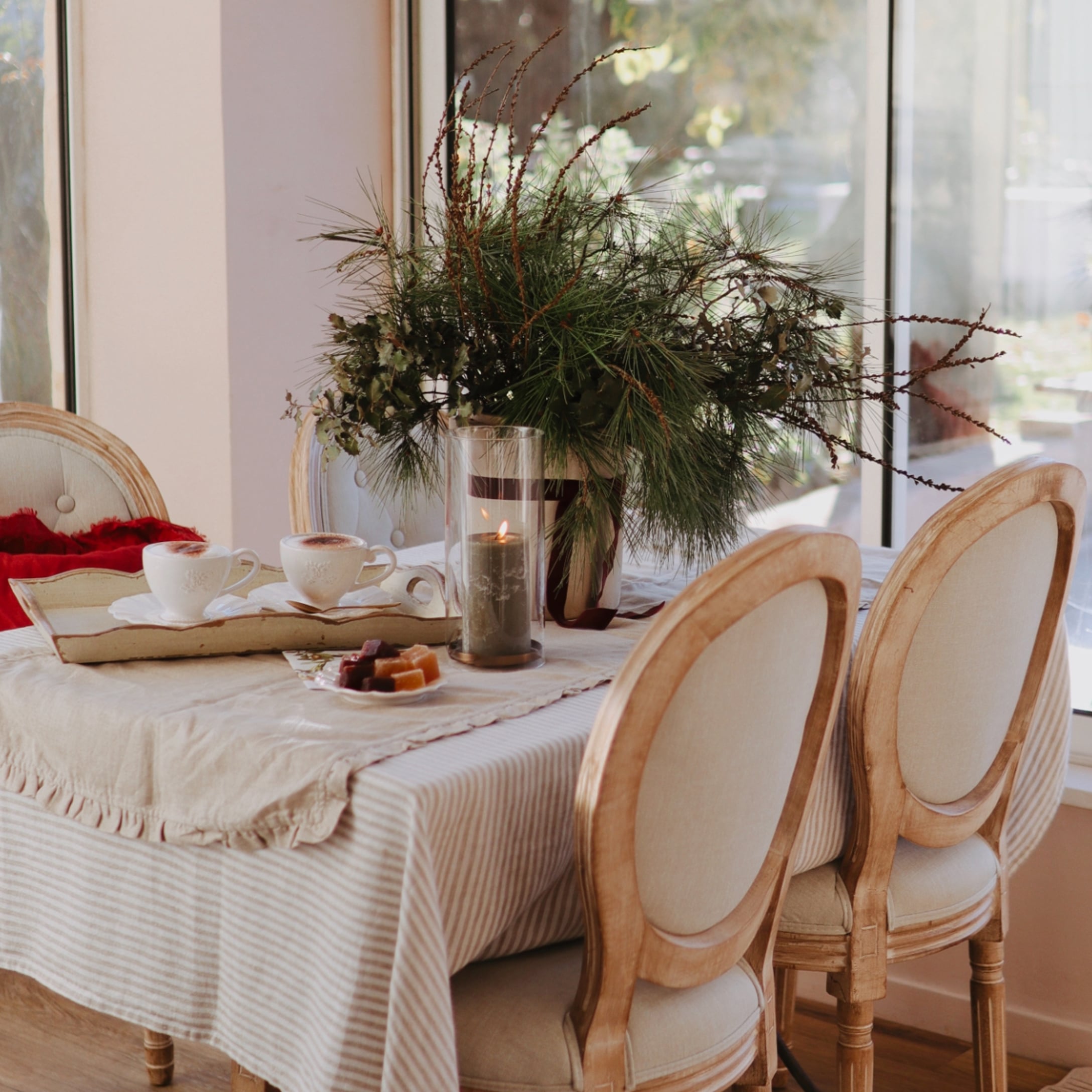 Tavolo con tovaglia beige, sedie in legno e decorazione natalizia con rami verdi e candela accesa