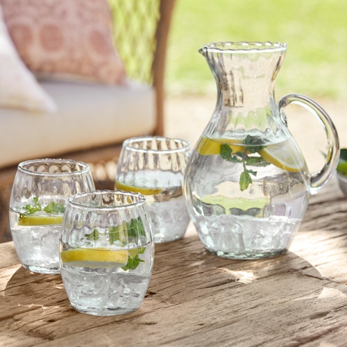 Caraffa di vetro e tre bicchieri con acqua, limone e menta su tavolo di legno all'aperto