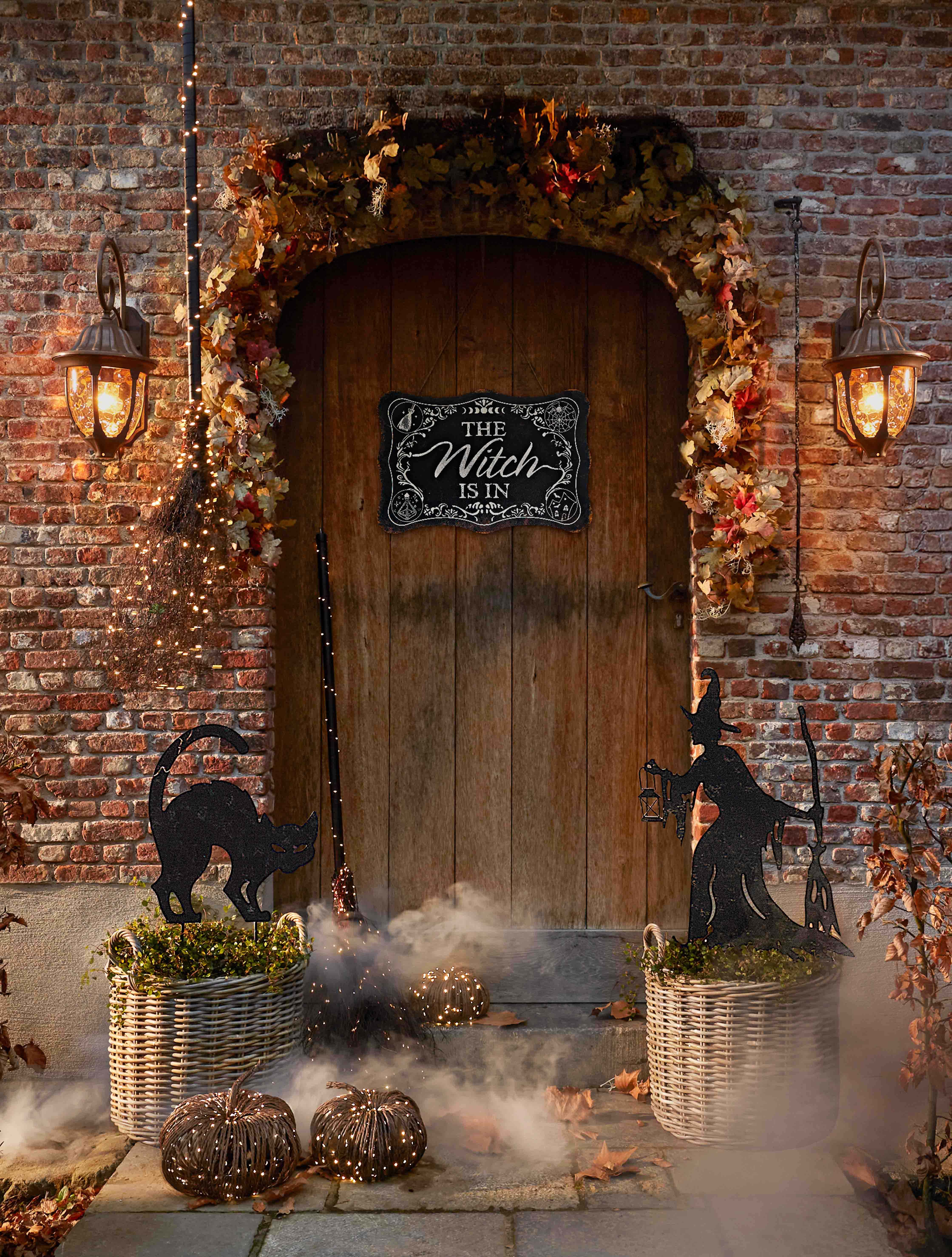 Decorazione di Halloween con strega, gatto nero e lanterne davanti a porta in legno e muro di mattoni