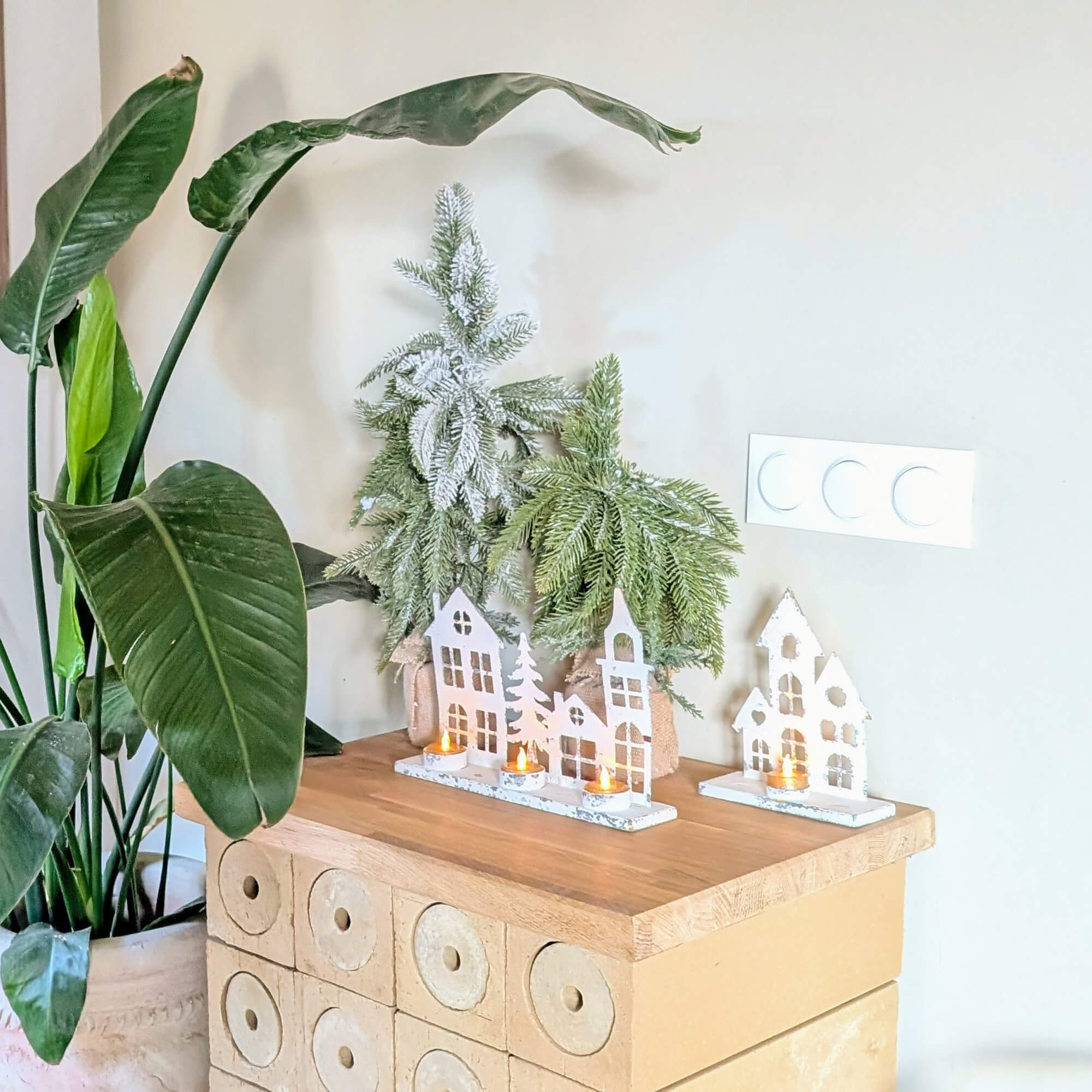 Decorazione natalizia con portacandele bianchi a forma di casette e rami verdi su piano in legno