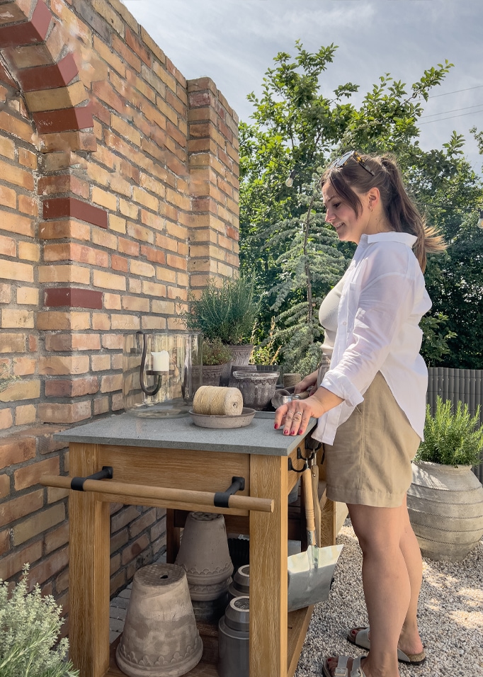 Tavolo da giardino in legno con piano in pietra e vasi, donna con camicia bianca e gonna beige