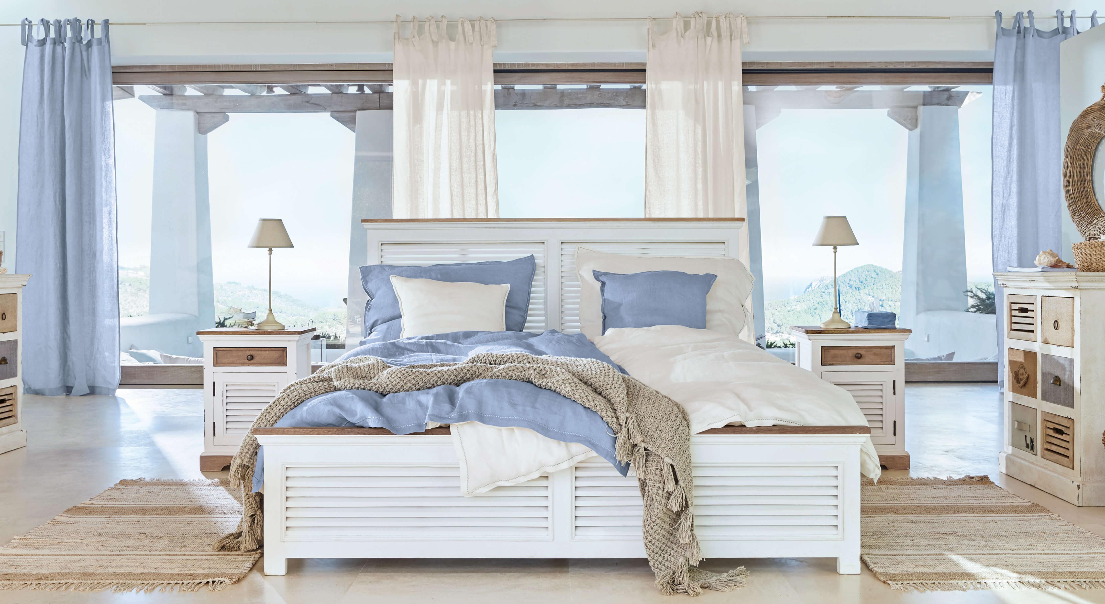 Letto in legno bianco con biancheria blu e beige, tappeto naturale in camera luminosa con vista