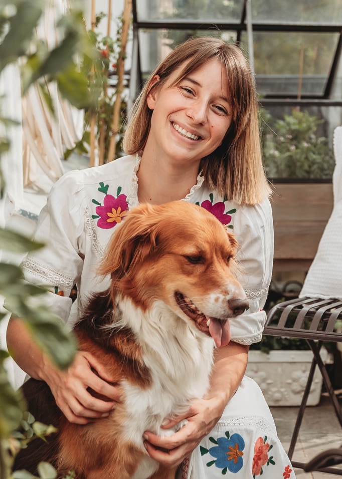 Donna con vestito bianco ricamato colorato che tiene un cane marrone e bianco in interno luminoso con piante