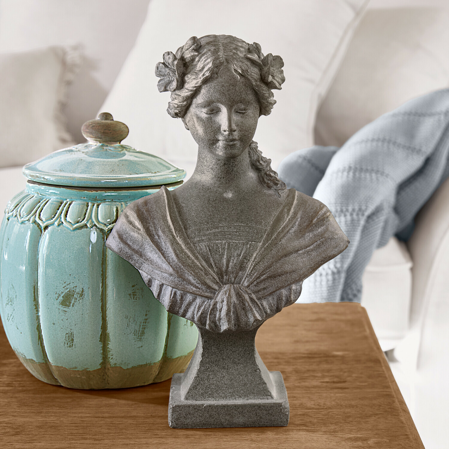 Busto femminile in pietra grigia con fiori tra i capelli su tavolo in legno accanto a vaso ceramico turchese