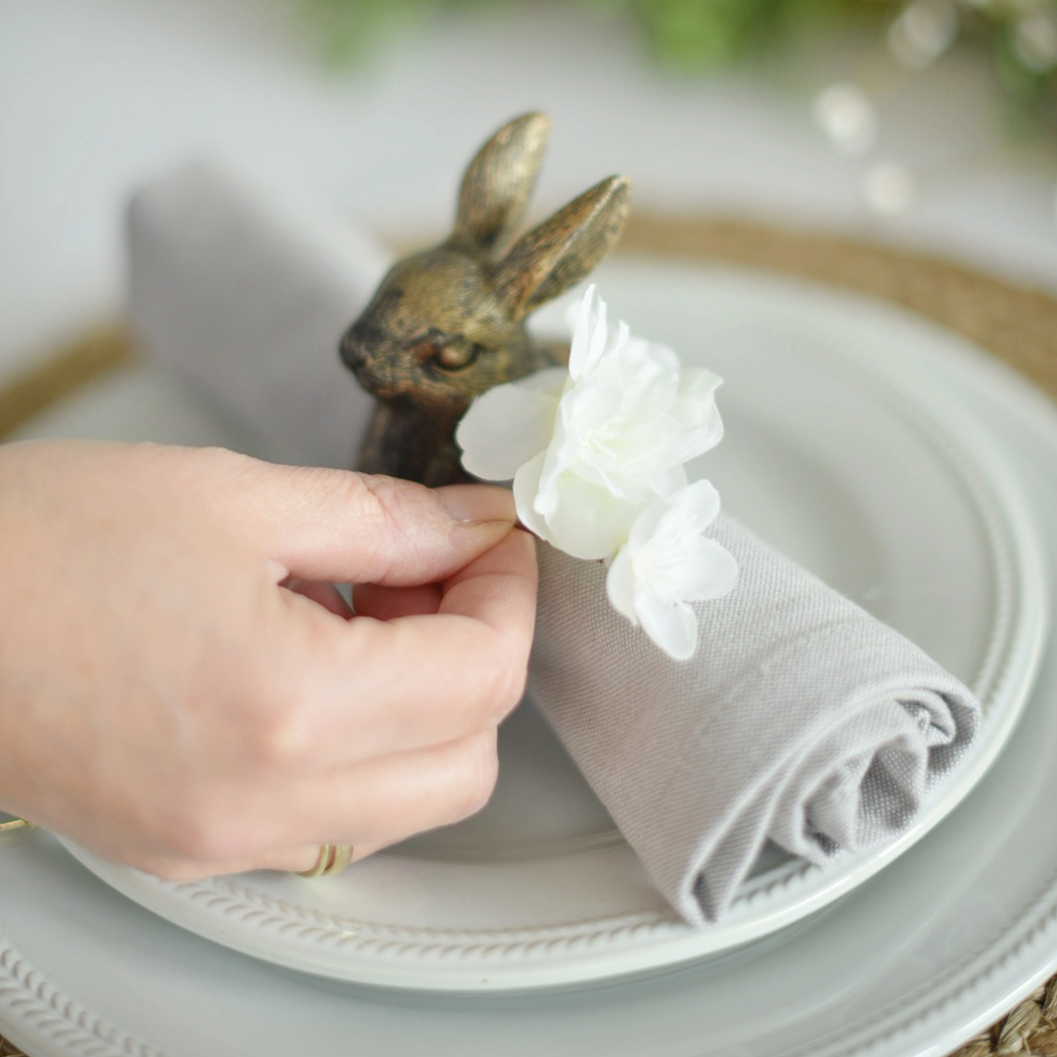 Anello portatovagliolo in lino grigio con fiore bianco e decorazione coniglio in ottone su piatto bianco
