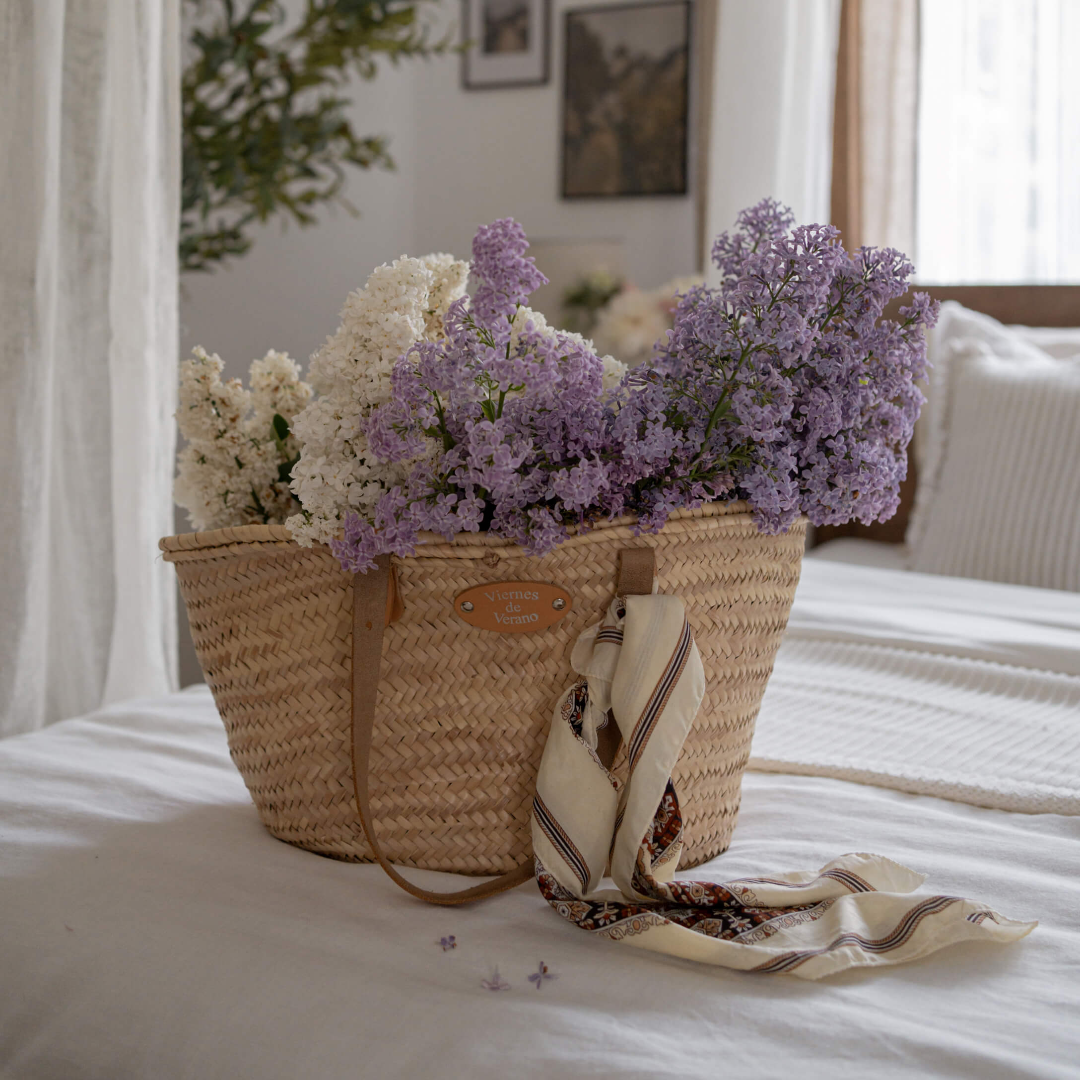 Cesto intrecciato con fiori bianchi e viola e foulard su letto bianco in stanza luminosa