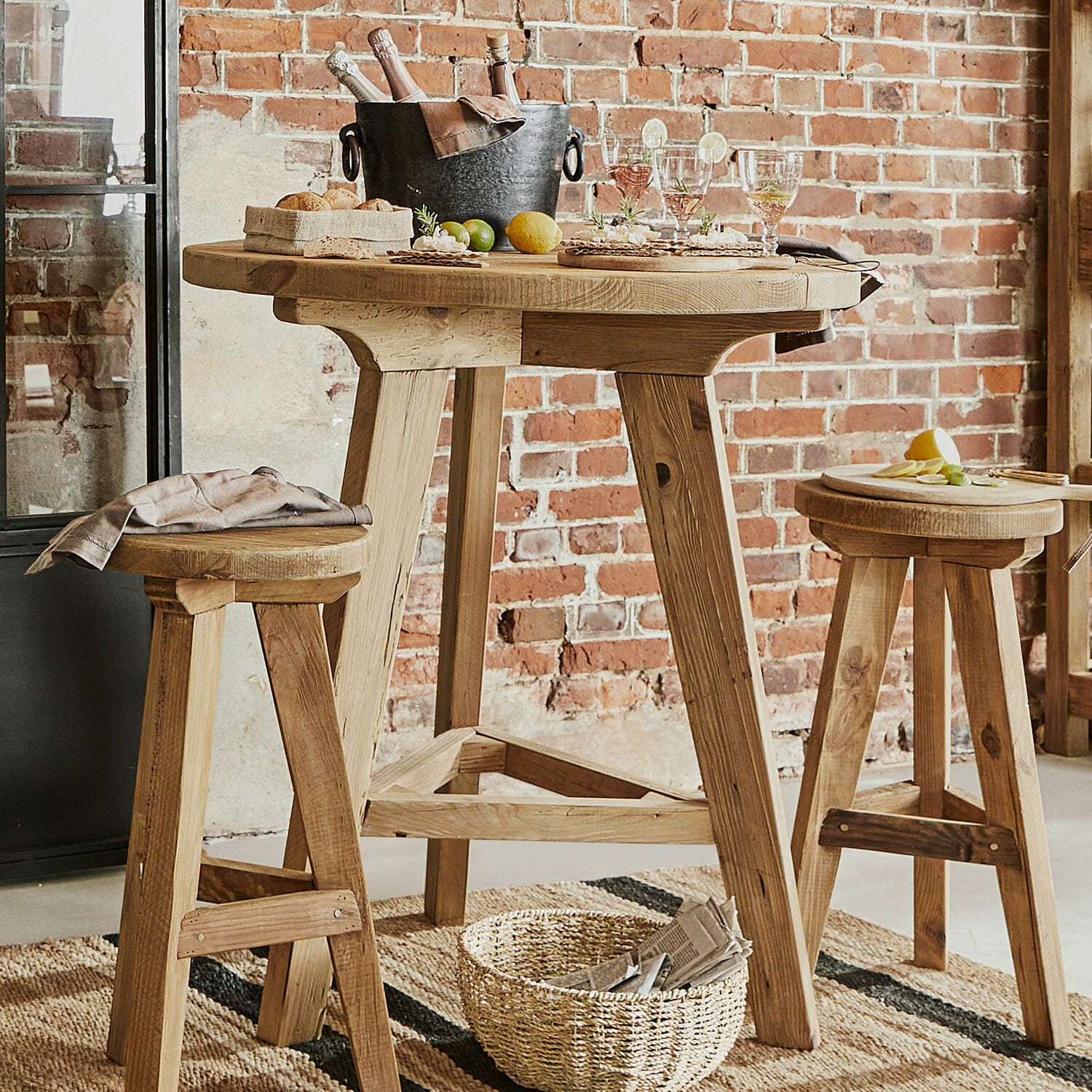Tavolino Stonington Tavolo alto in legno naturale con due sgabelli davanti a muro di mattoni, stile loft