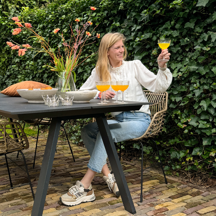Donna con camicetta bianca e jeans seduta a tavolo nero all'aperto, tiene bicchiere con bevanda arancione, vaso con fiori accanto