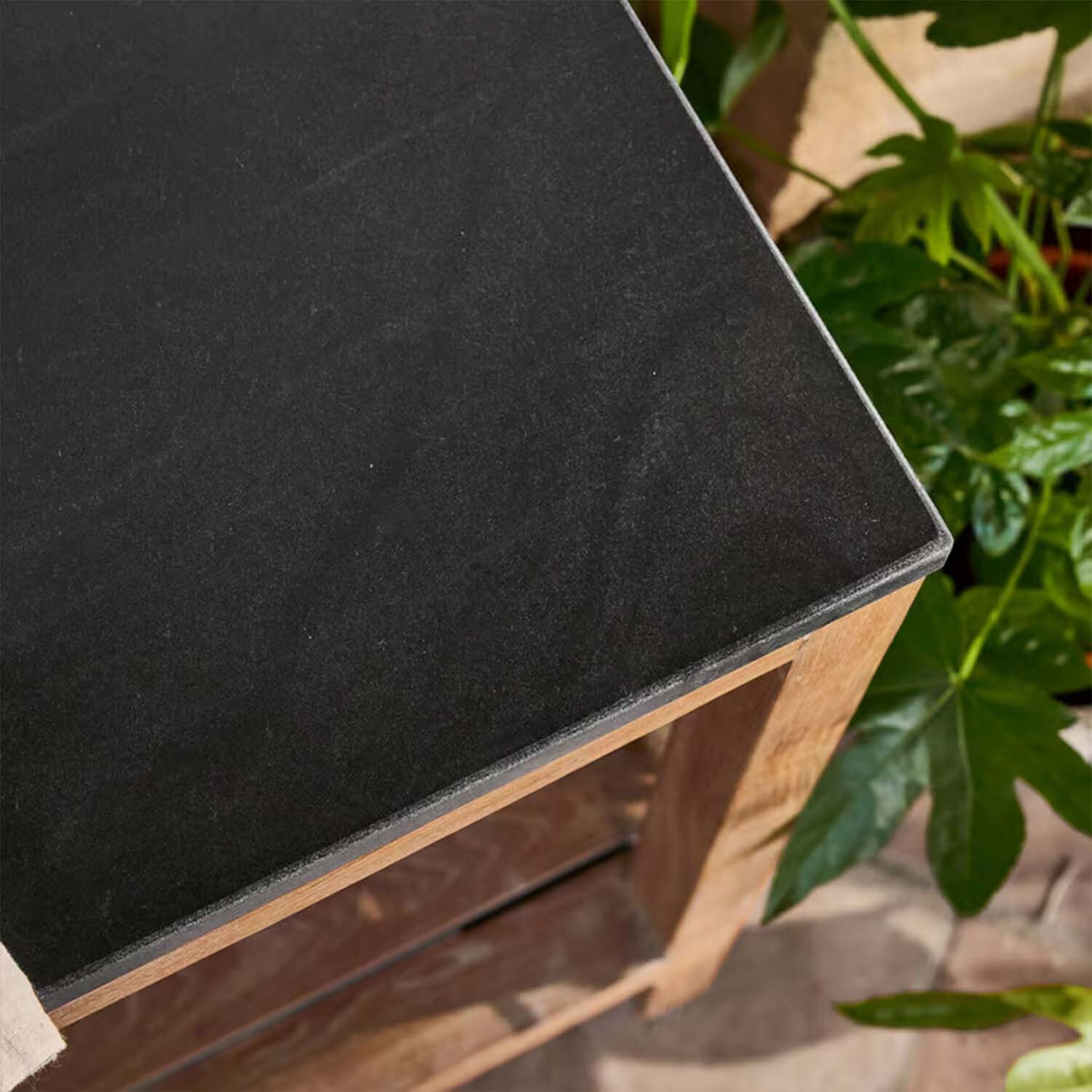 Piano tavolo in pietra nera con base in legno circondato da piante verdi, arredo da giardino