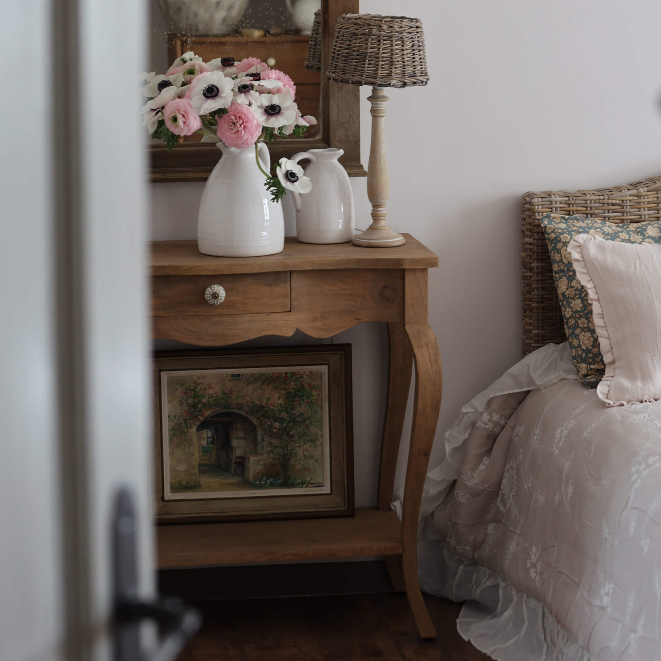 Tavolino in legno con brocche in ceramica bianca e fiori accanto a letto con testiera in vimini