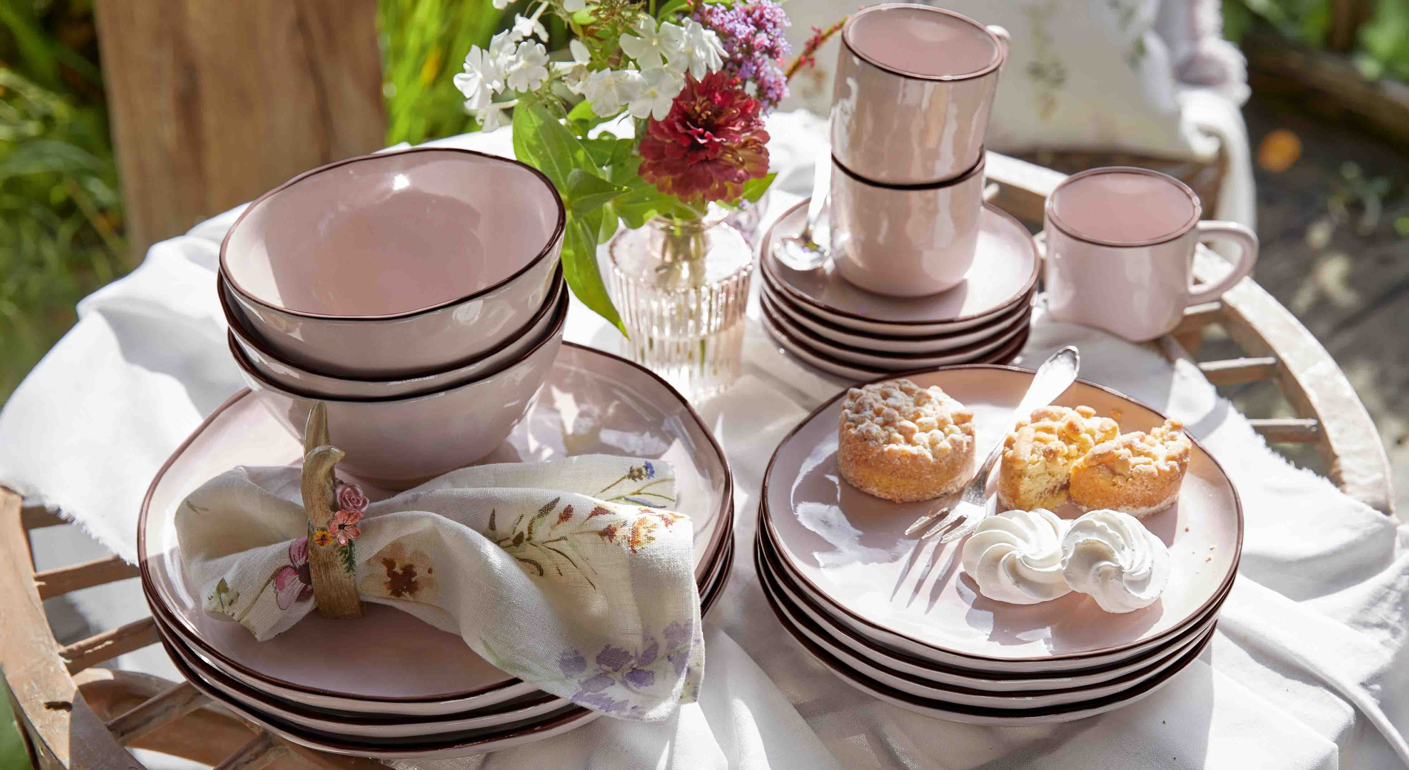 Set di stoviglie in ceramica rosa con piatti, ciotole e tazze su tavolo in legno con dolci e fiori