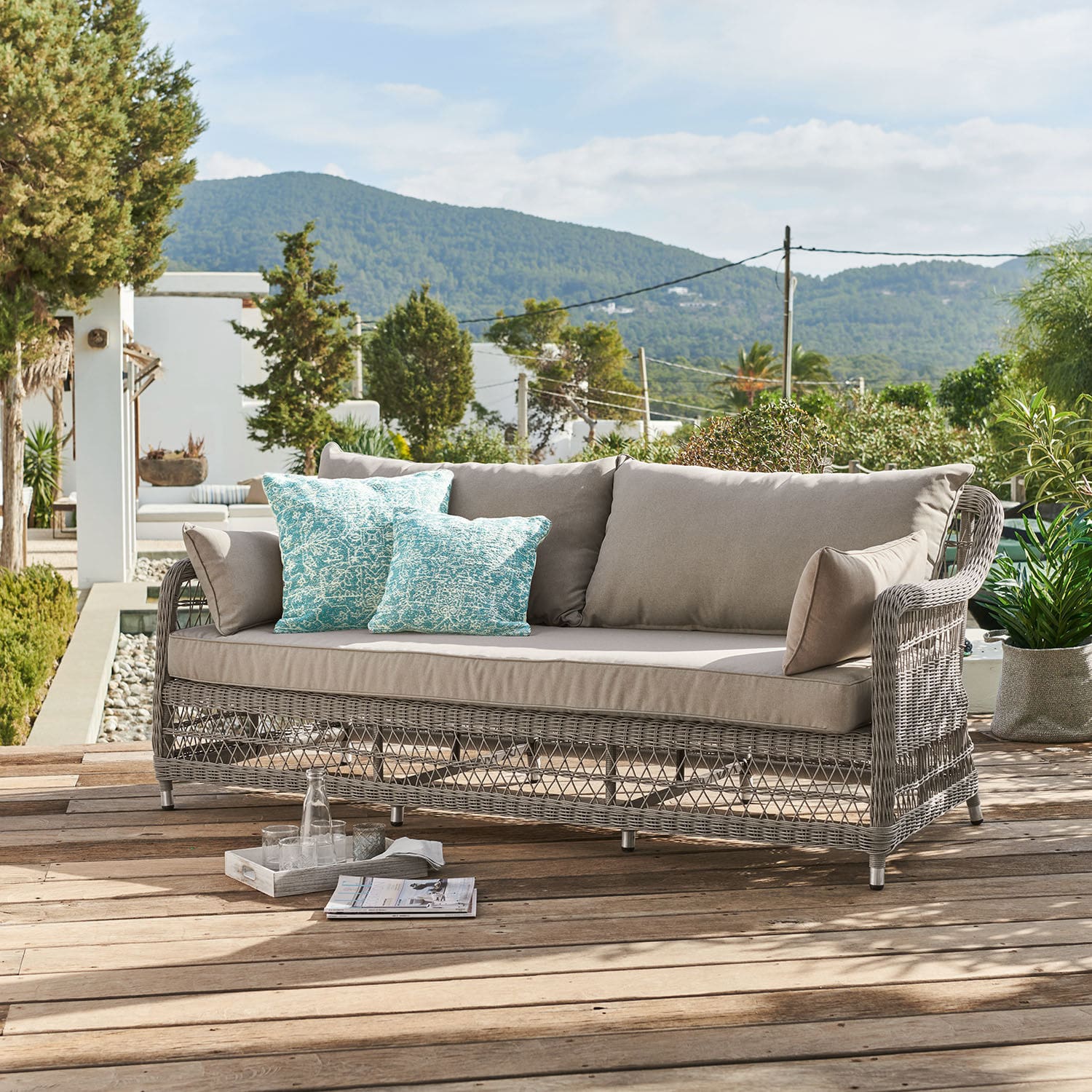 Divano Tanaiste Divano da giardino in rattan grigio con cuscini beige e cuscini decorativi turchesi su terrazza in legno