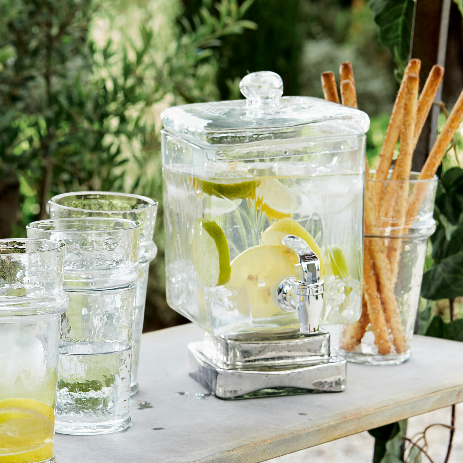 Dispenser di bevande Logan Dispenser per bevande in vetro con rubinetto, pieno di acqua al limone, su tavolo con bicchieri e grissini