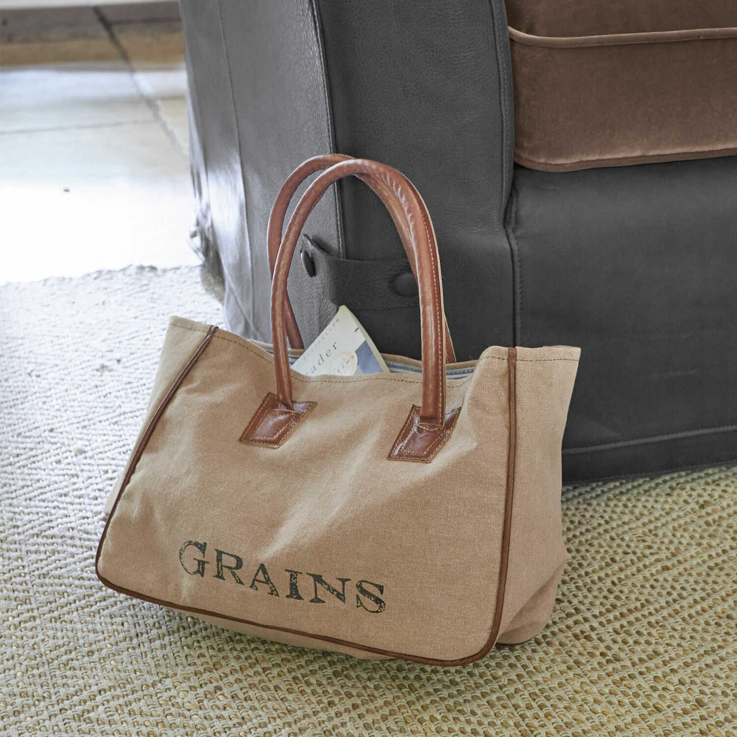 Borsa Grains Borsa beige in tessuto con manici in pelle marrone e scritta GRAINS su tappeto vicino al divano
