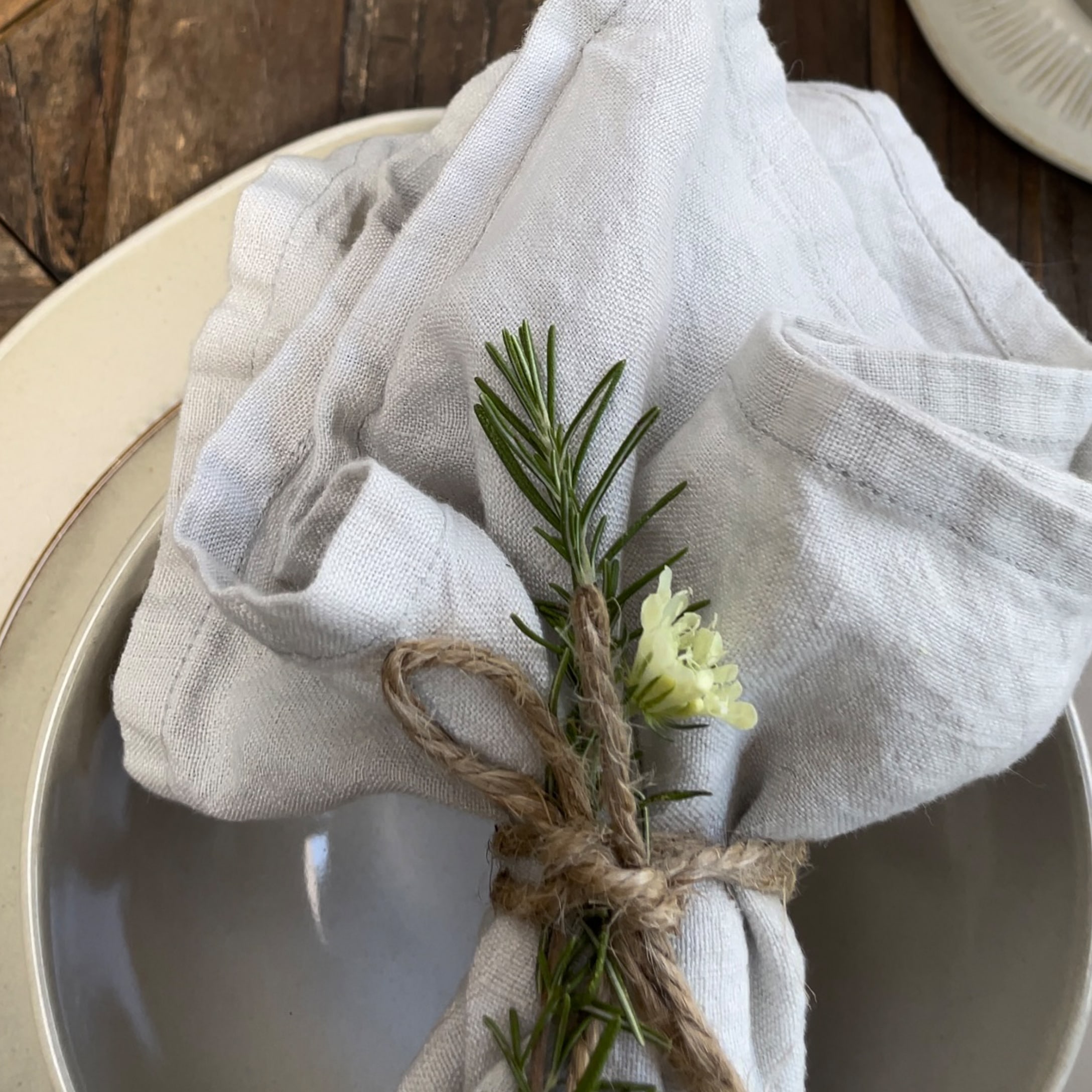 Tovagliolo di lino grigio chiaro con rametto di rosmarino e fiore su piatto, stile rustico