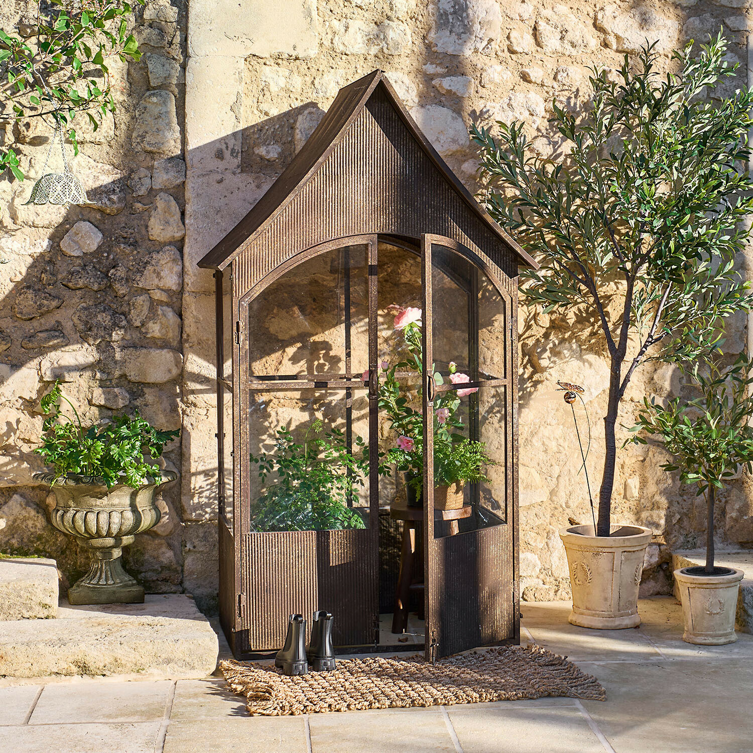 Serra da giardino in metallo marrone stile vintage con piante e vasi davanti a muro di pietra soleggiato