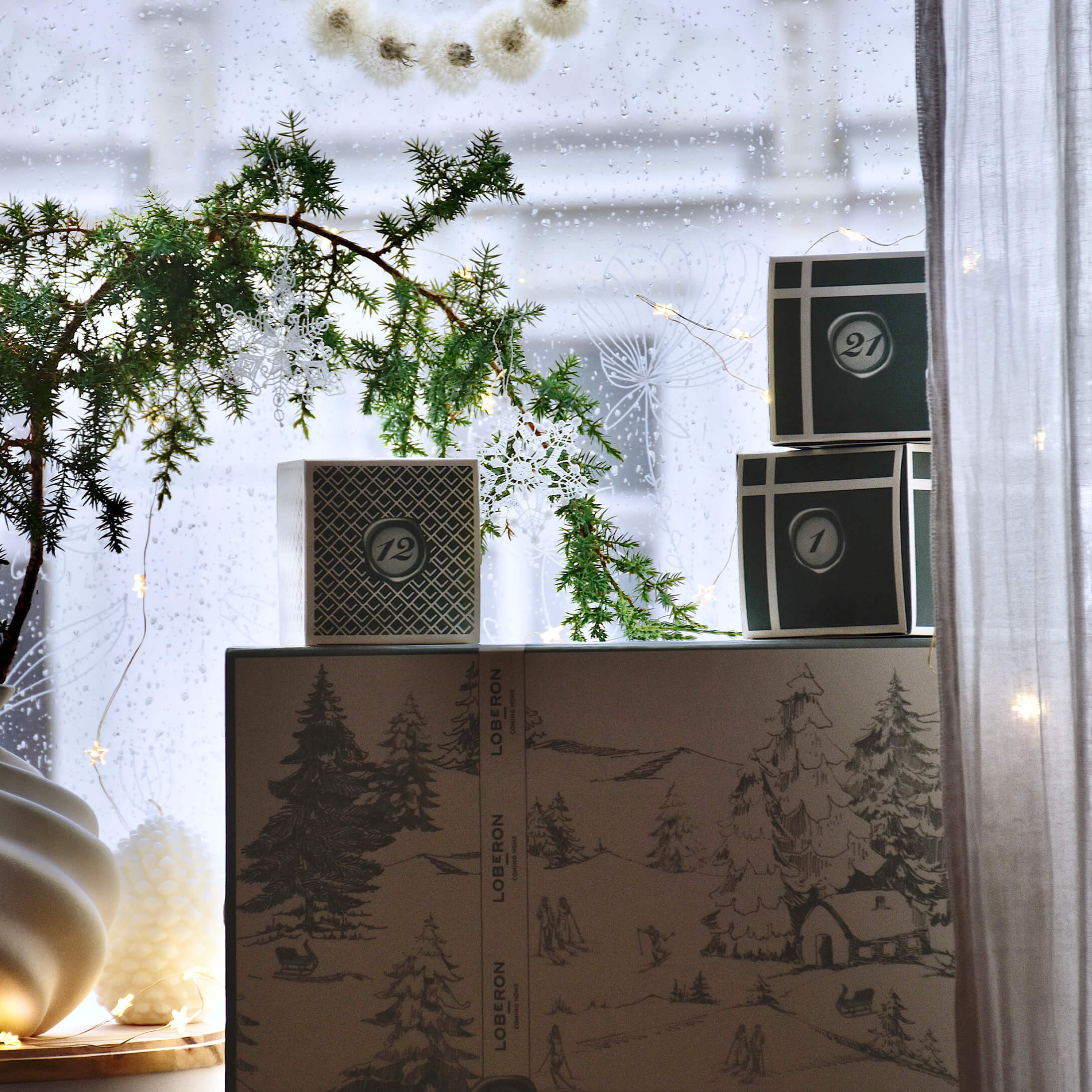 Scatole decorative con motivo invernale e ramo verde con fiocchi bianchi davanti a finestra piovosa