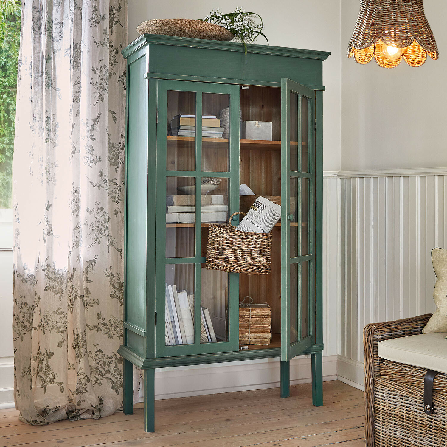 Mobile alto Faymere Vetrina in legno verde con ante in vetro e cesta in vimini in soggiorno luminoso con luce naturale