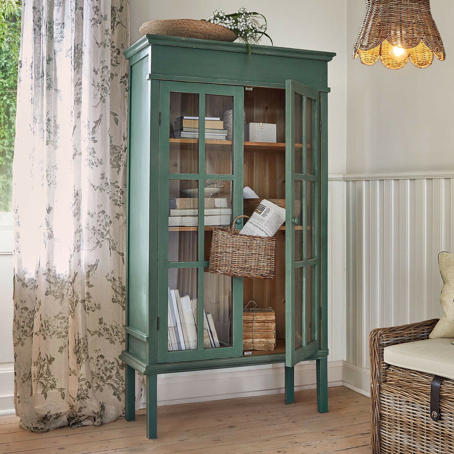 Mobile alto Faymere Vetrina in legno verde con ante in vetro e cesta in vimini in soggiorno luminoso con luce naturale