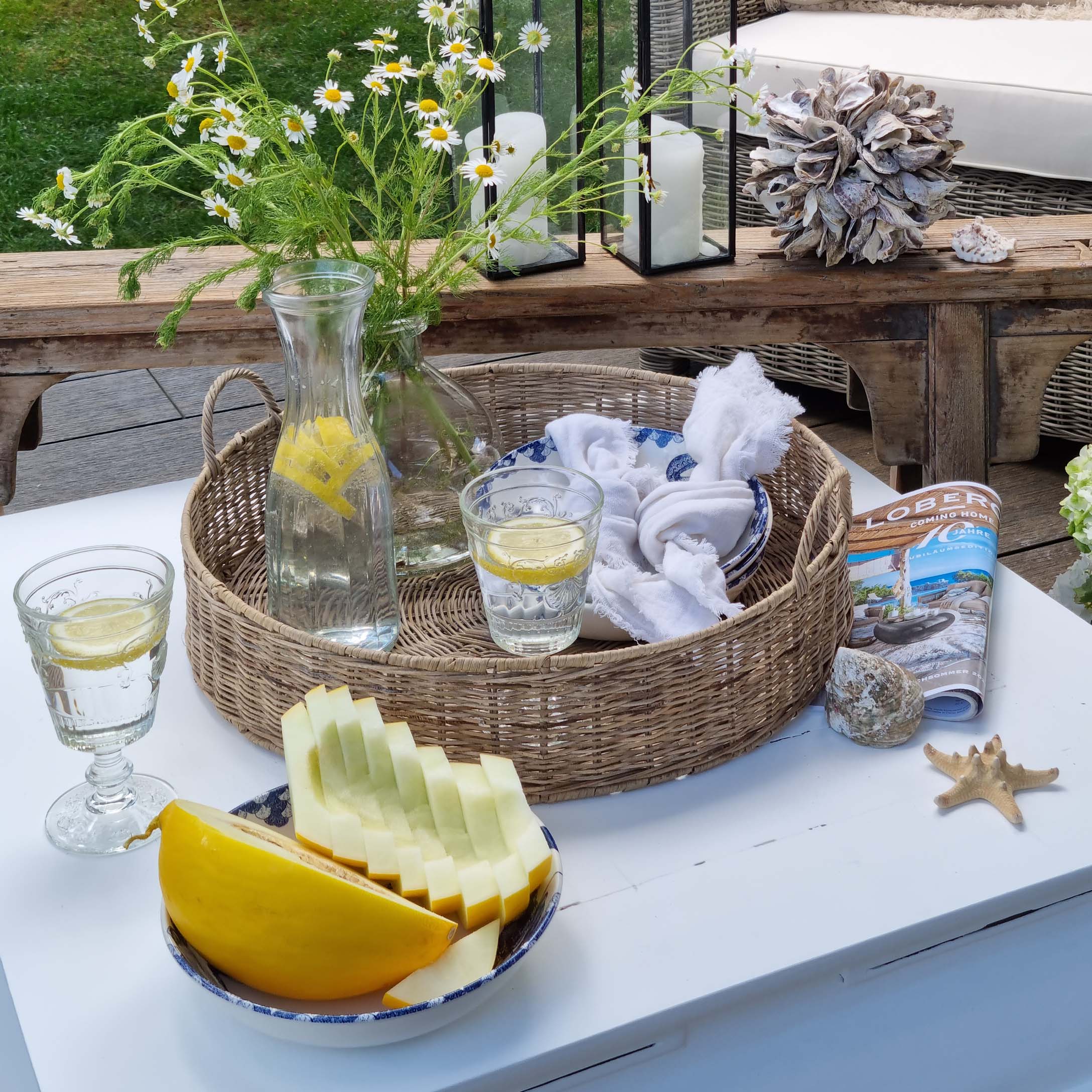 Cesto rotondo in vimini con caraffa d'acqua al limone e bicchiere su tavolo bianco in giardino