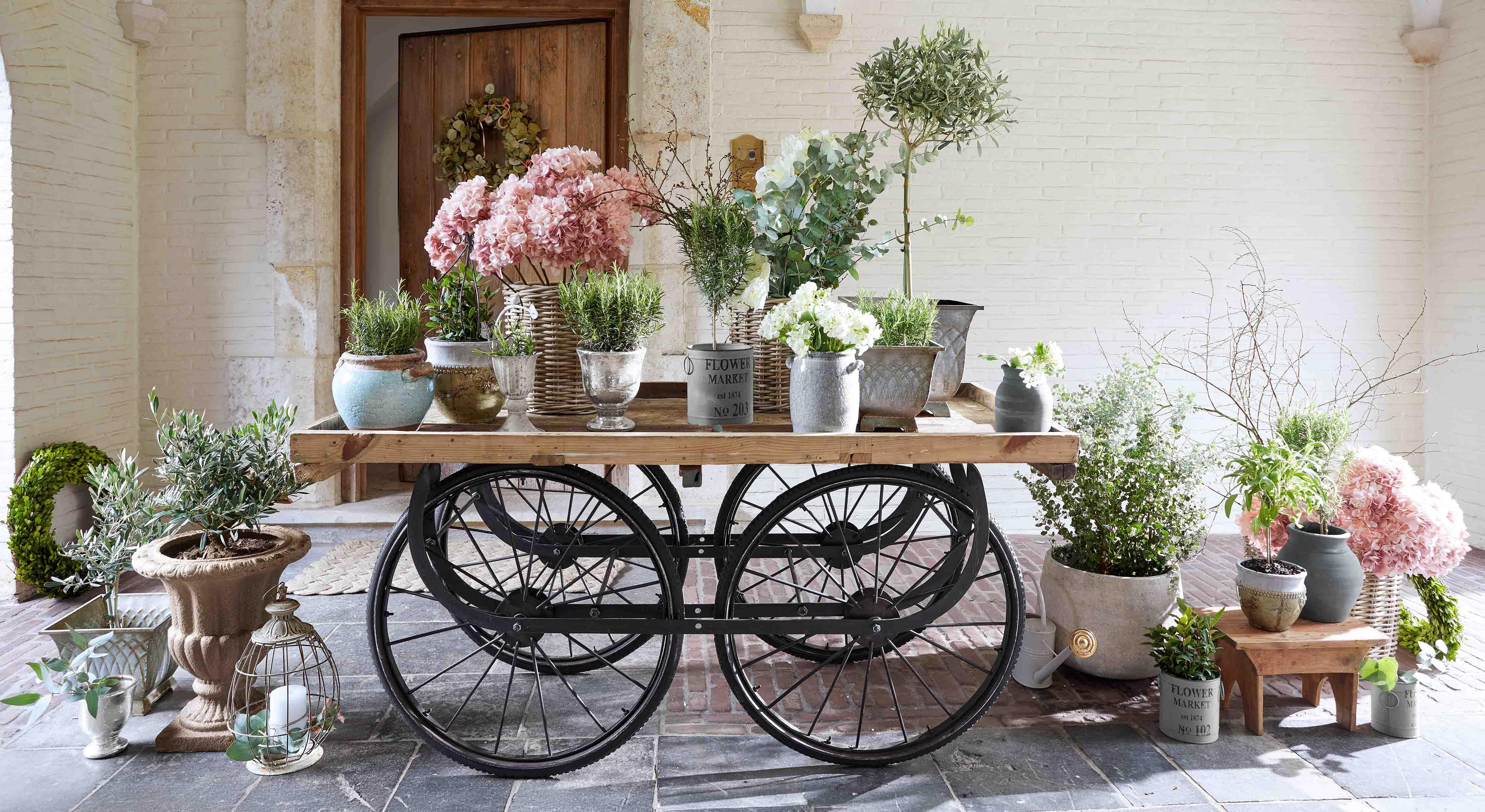 Carrello in legno con ruote nere e vasi con fiori rosa e bianchi di varie dimensioni